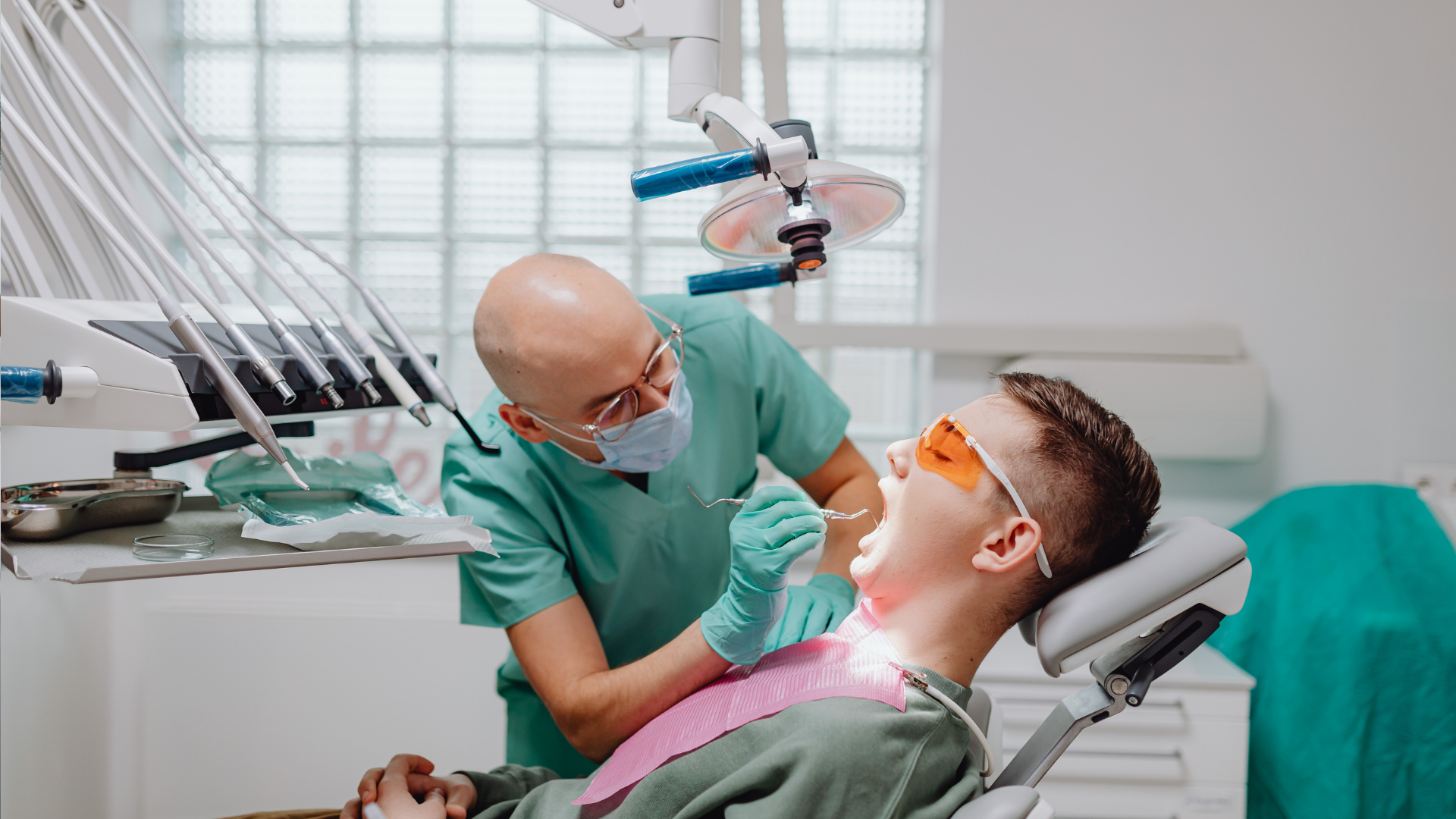 Dentist examining patient's teeth with tools in a dental office. Patient wears orange safety glasses.