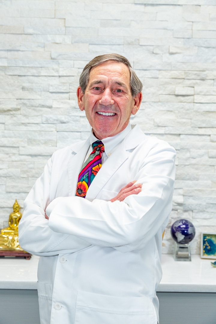 A man in a lab coat and tie is smiling in front of a framed diploma