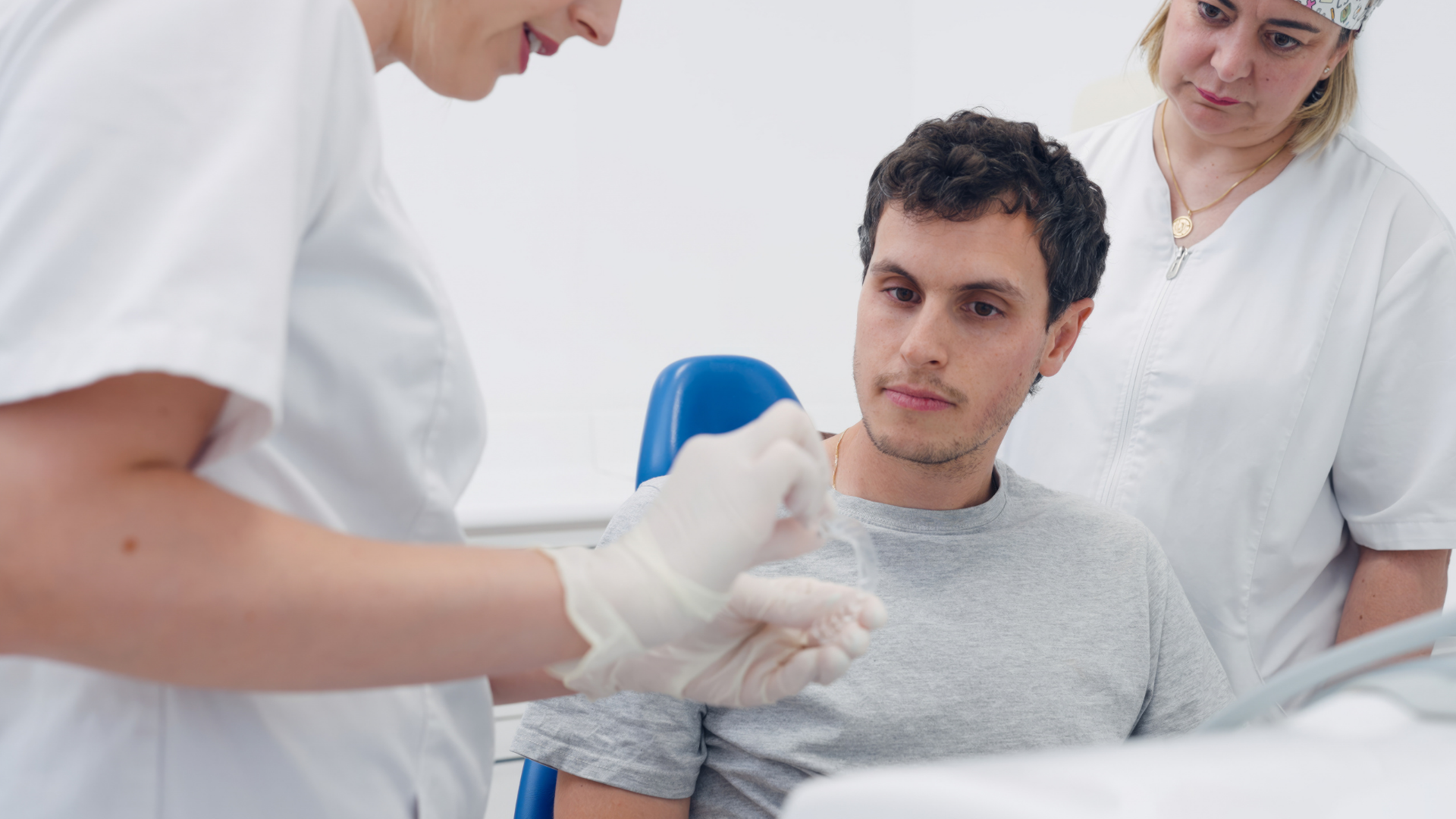 A patient in a gray shirt sits in a dental chair while two healthcare workers in white uniforms prepare tools.