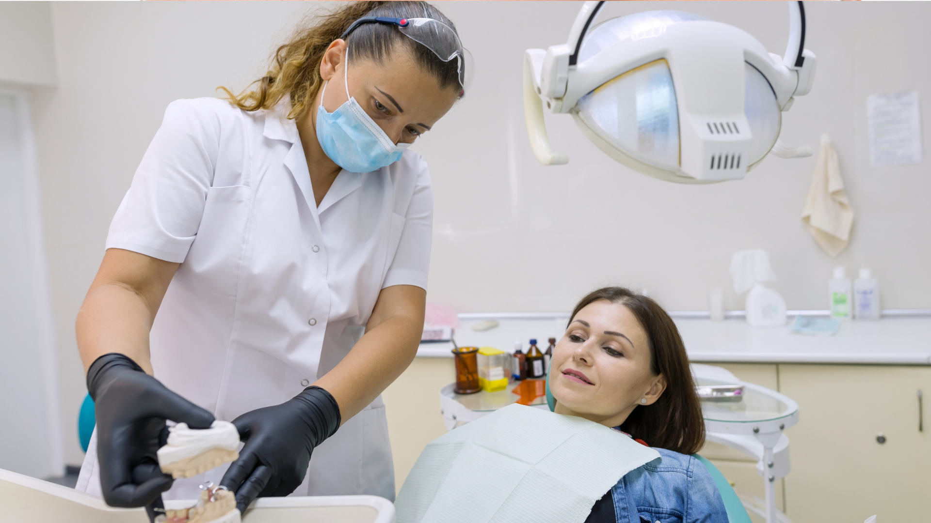 Dentist in white uniform and mask treating a patient in a dental chair, with overhead exam light.
