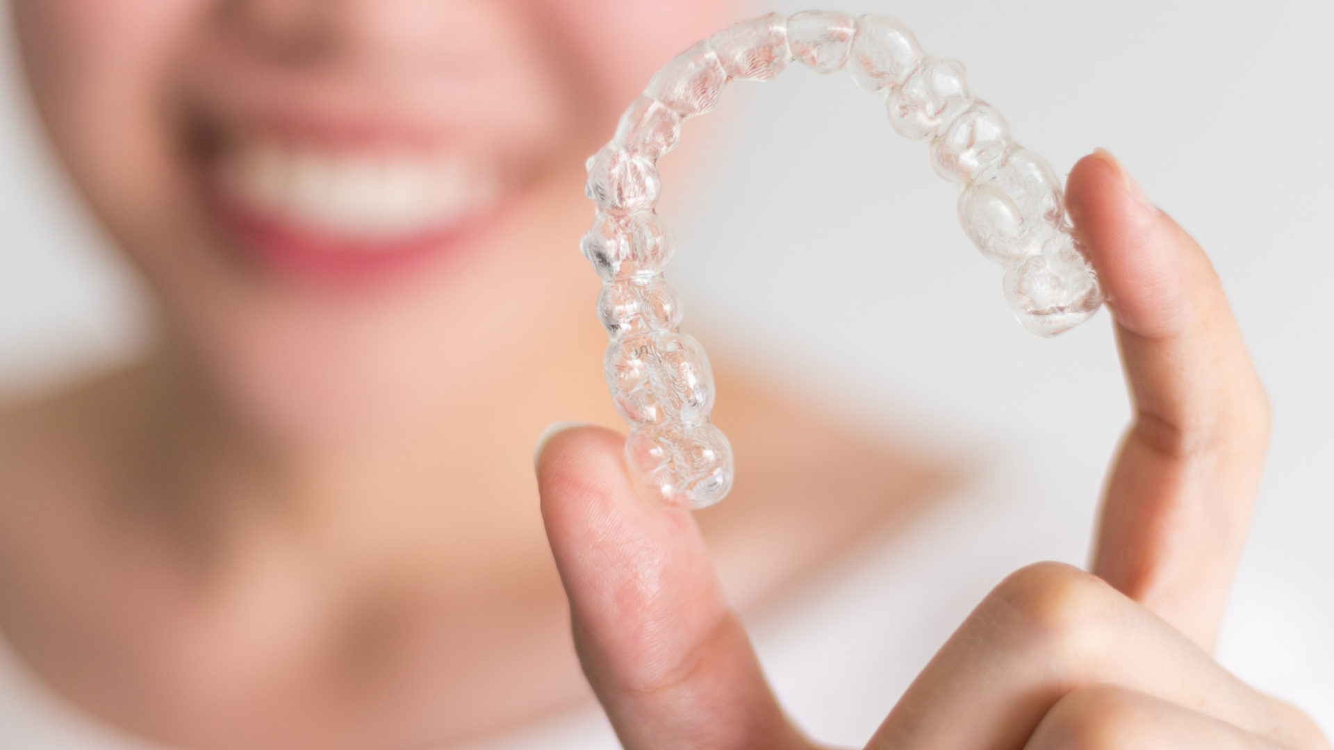 Hand holding a clear dental aligner in front of a smiling person’s mouth
