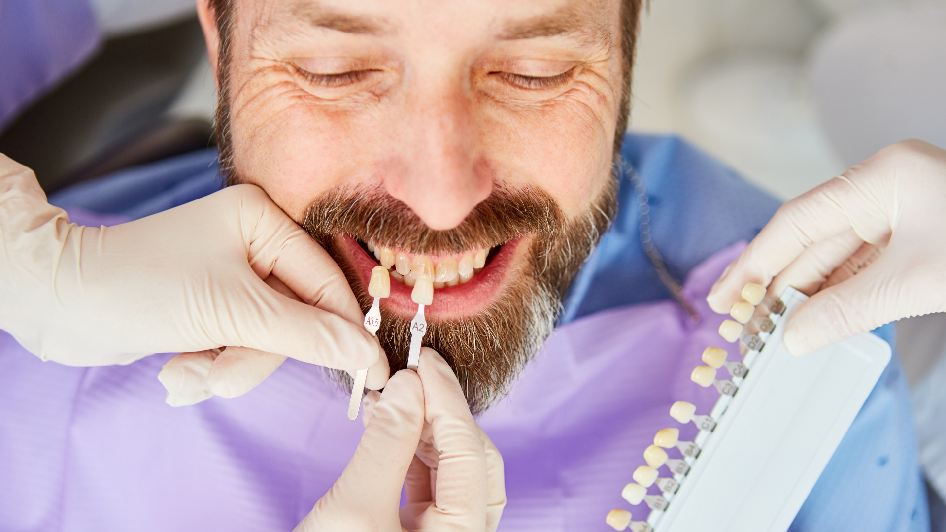 Man at dentist selecting tooth shade with a shade guide, hands in gloves, smiling.