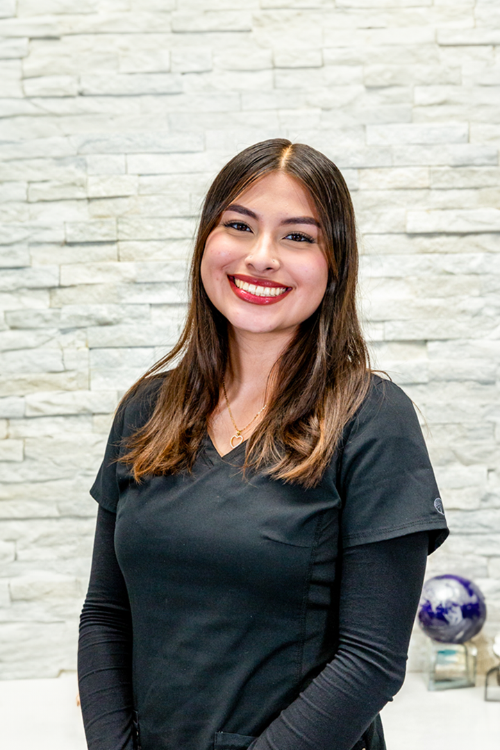 A woman in a black scrub top is smiling in front of a brick wall.