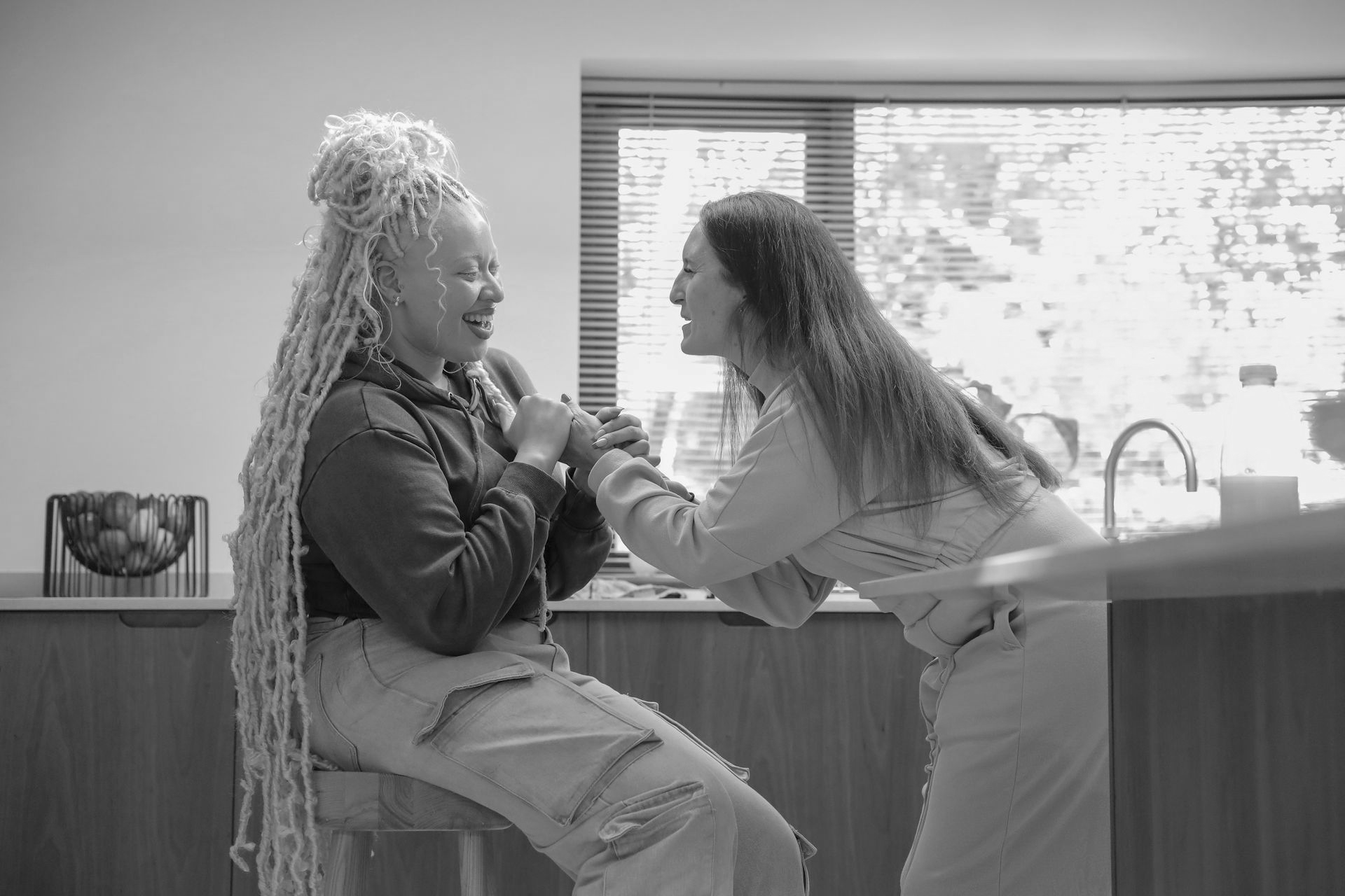 Two people smiling, holding hands. One with long dreadlocks seated, other leaning in near a window.