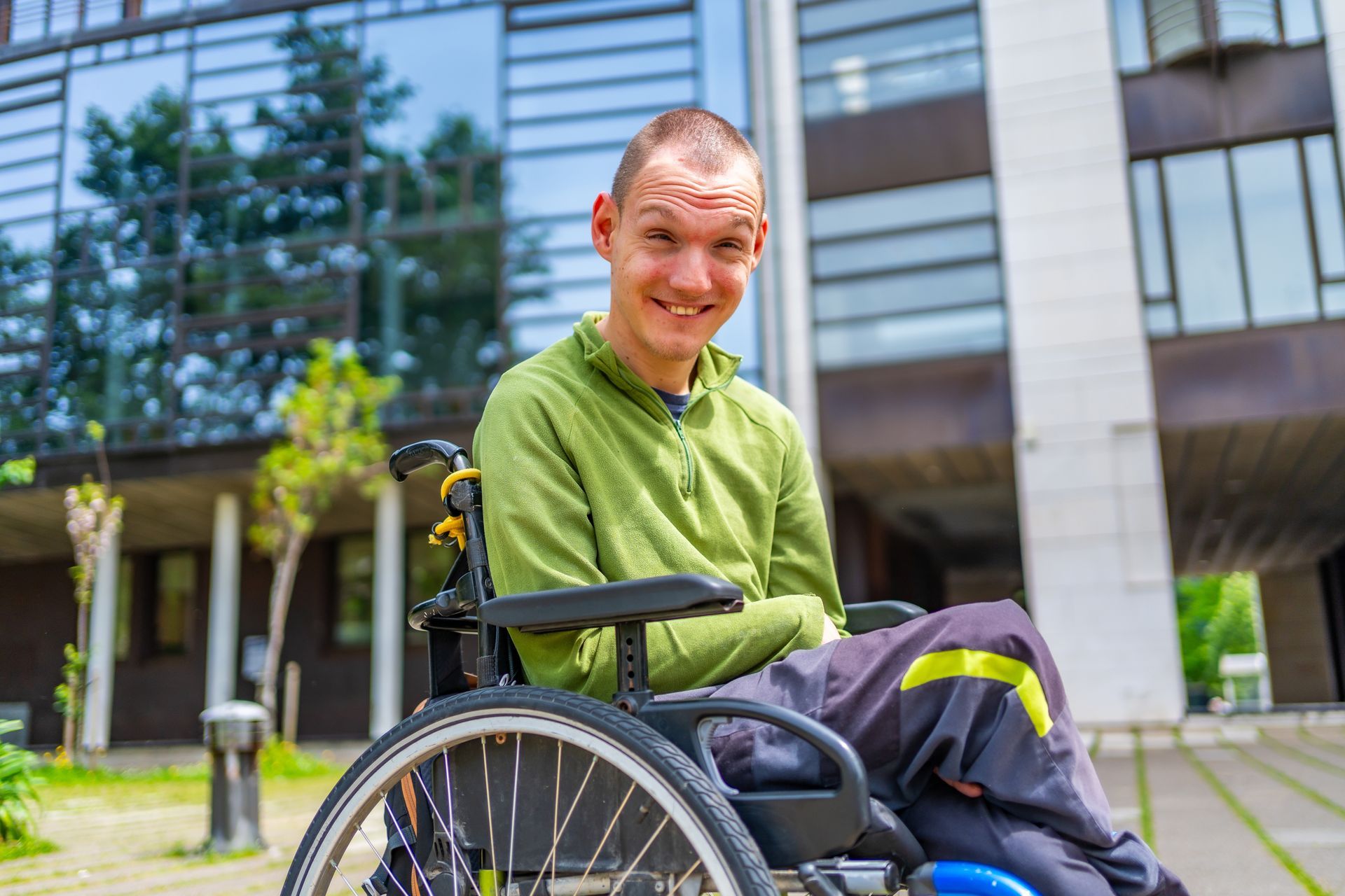 Smiling person in a wheelchair outdoors in front of a modern building.