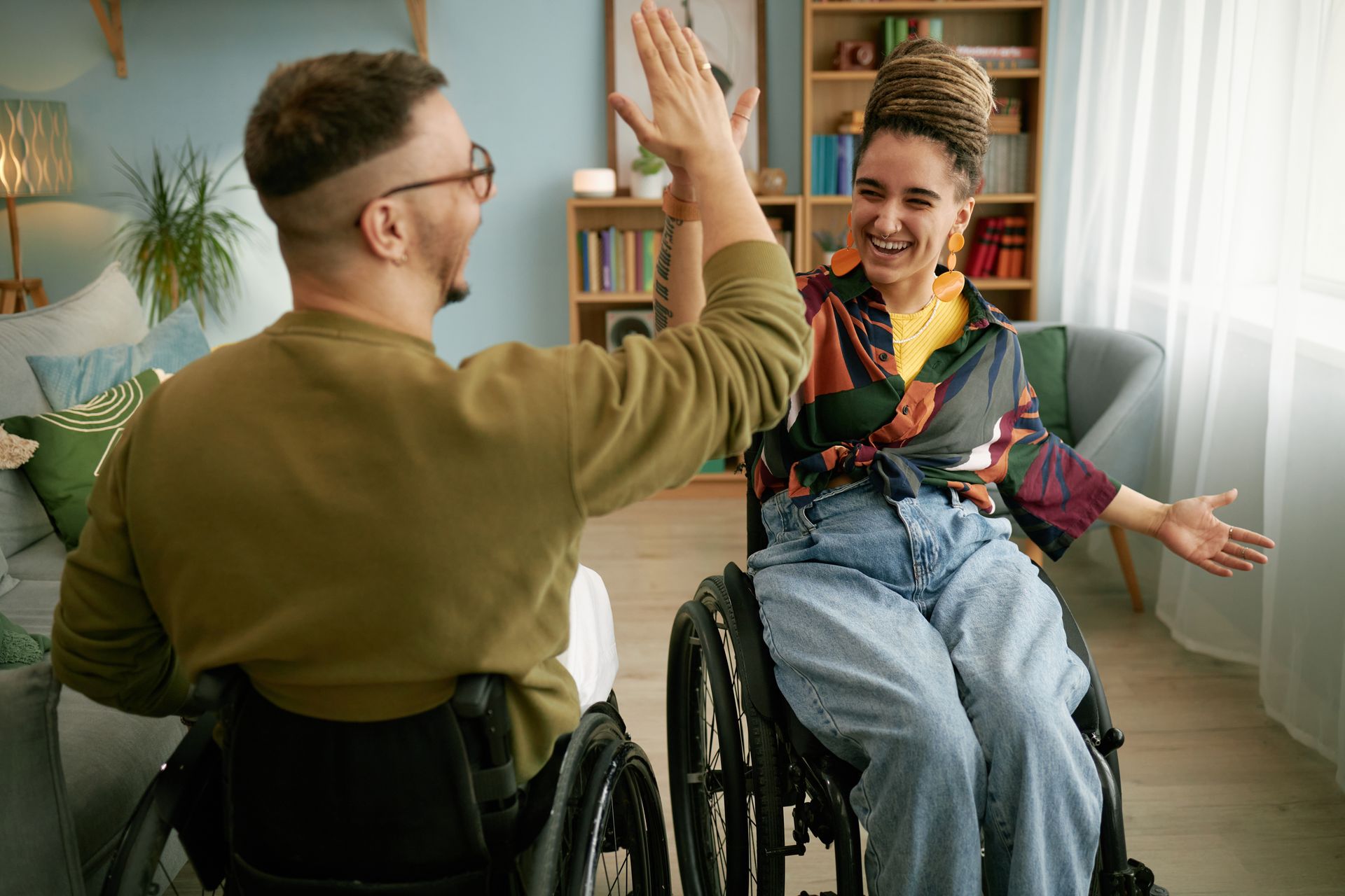 Two people in wheelchairs high-fiving in a well-lit living room, smiling.