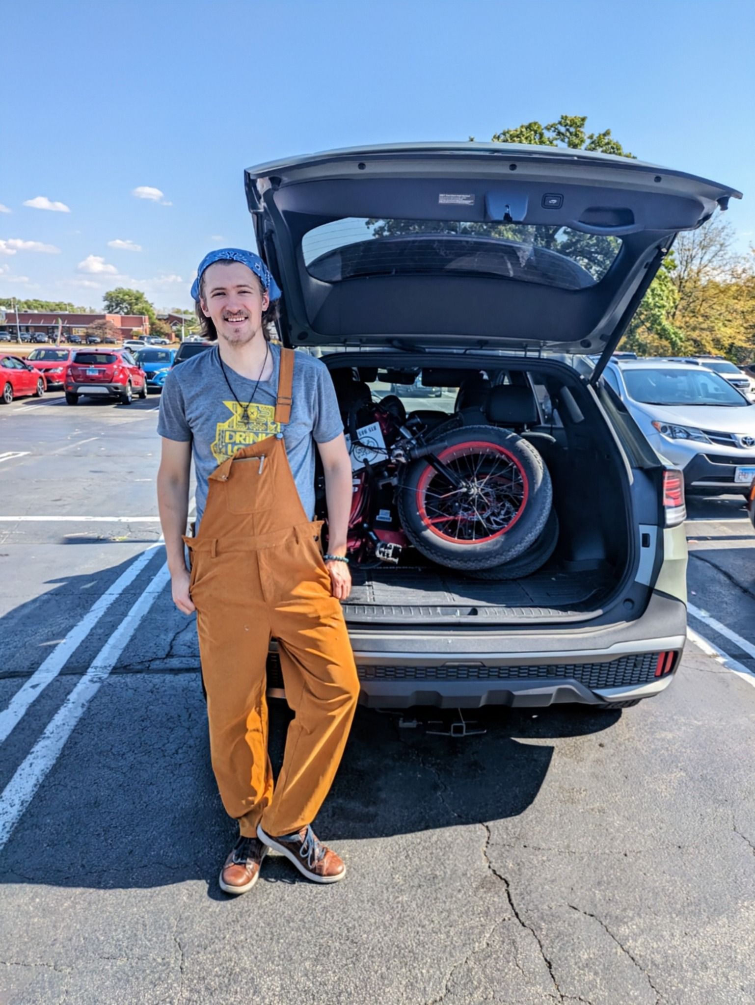 Person in orange overalls leans against a car with its hatch open, showing a bicycle in the trunk.