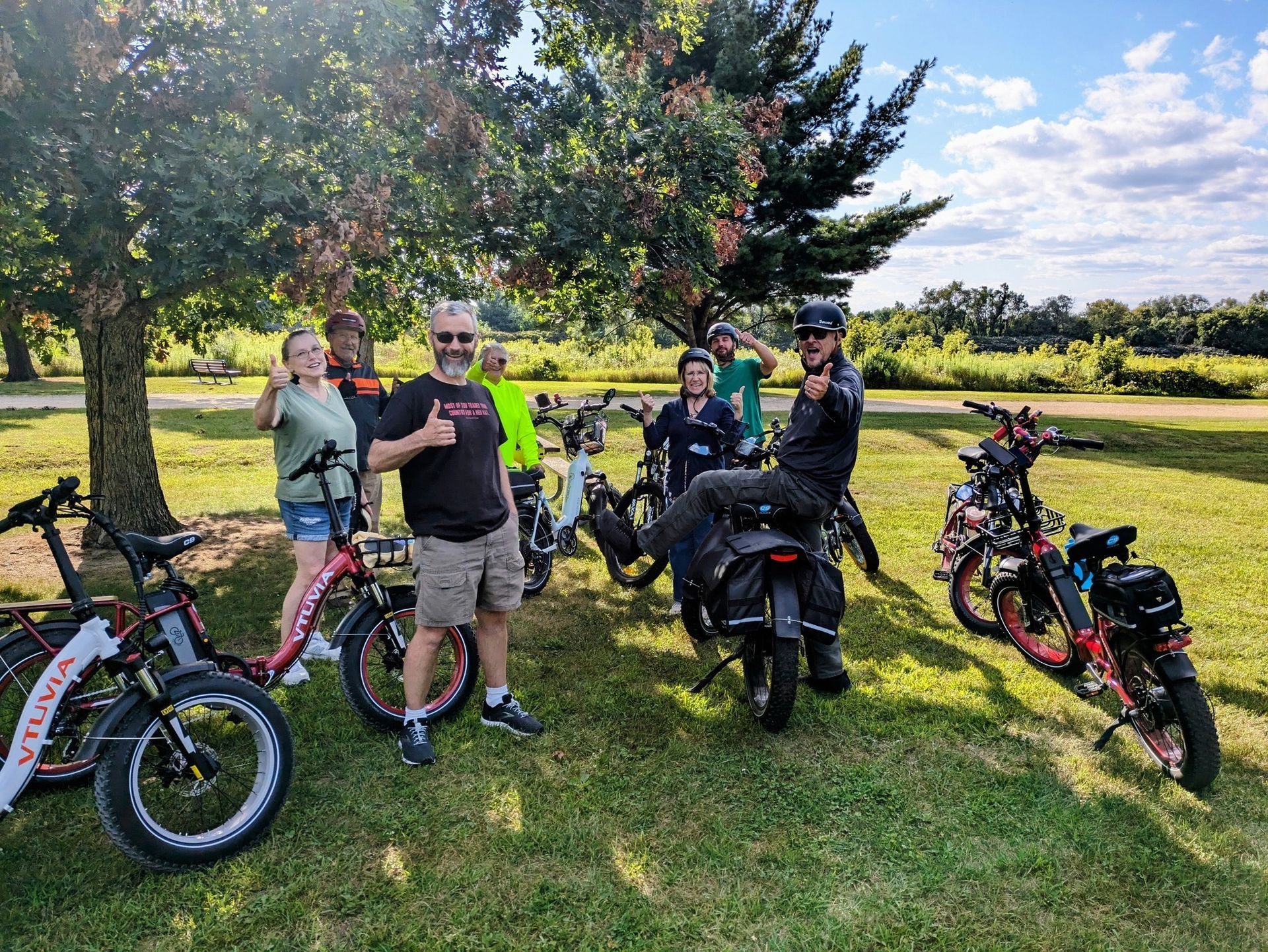 Cyclists posing with bikes on a grassy field beside trees under a sunny sky