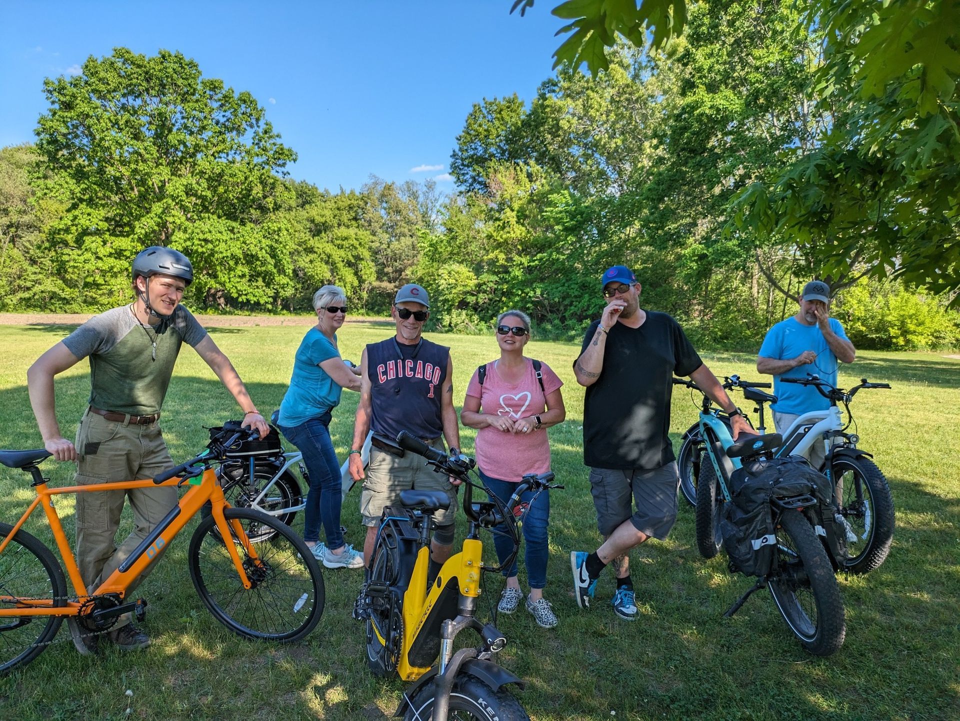 Cyclists posing with bikes on a sunny grassy trail, trees and blue sky in the background.