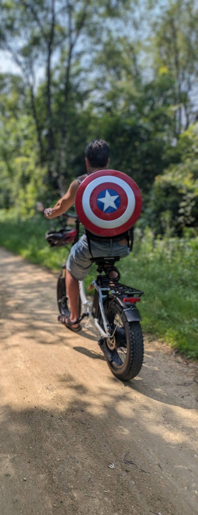 Person rides a bicycle on a dirt path with a Captain America shield on their back.