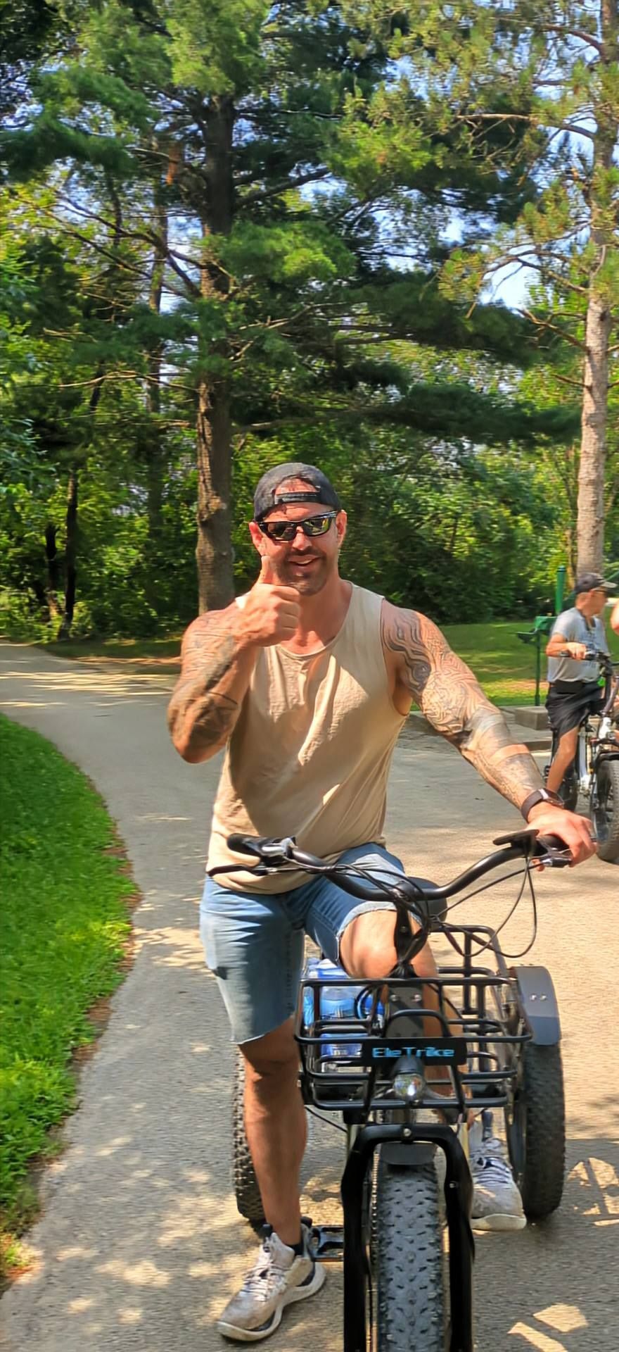 Man on a bicycle on a sunny park path, smiling and holding a drink, with trees and another cyclist behind him