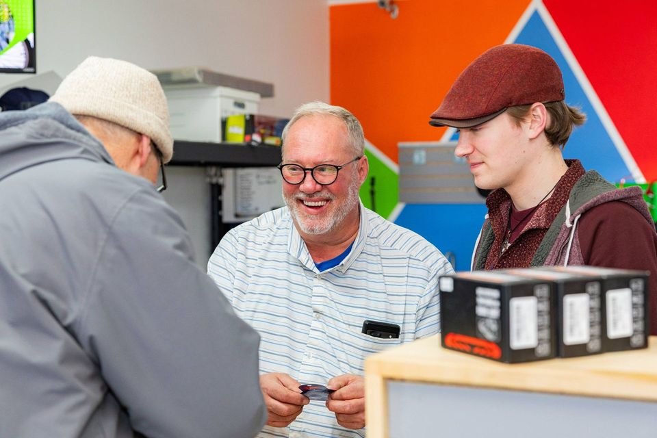 Three people chatting at a table in a colorful workshop, with boxes and tools nearby.