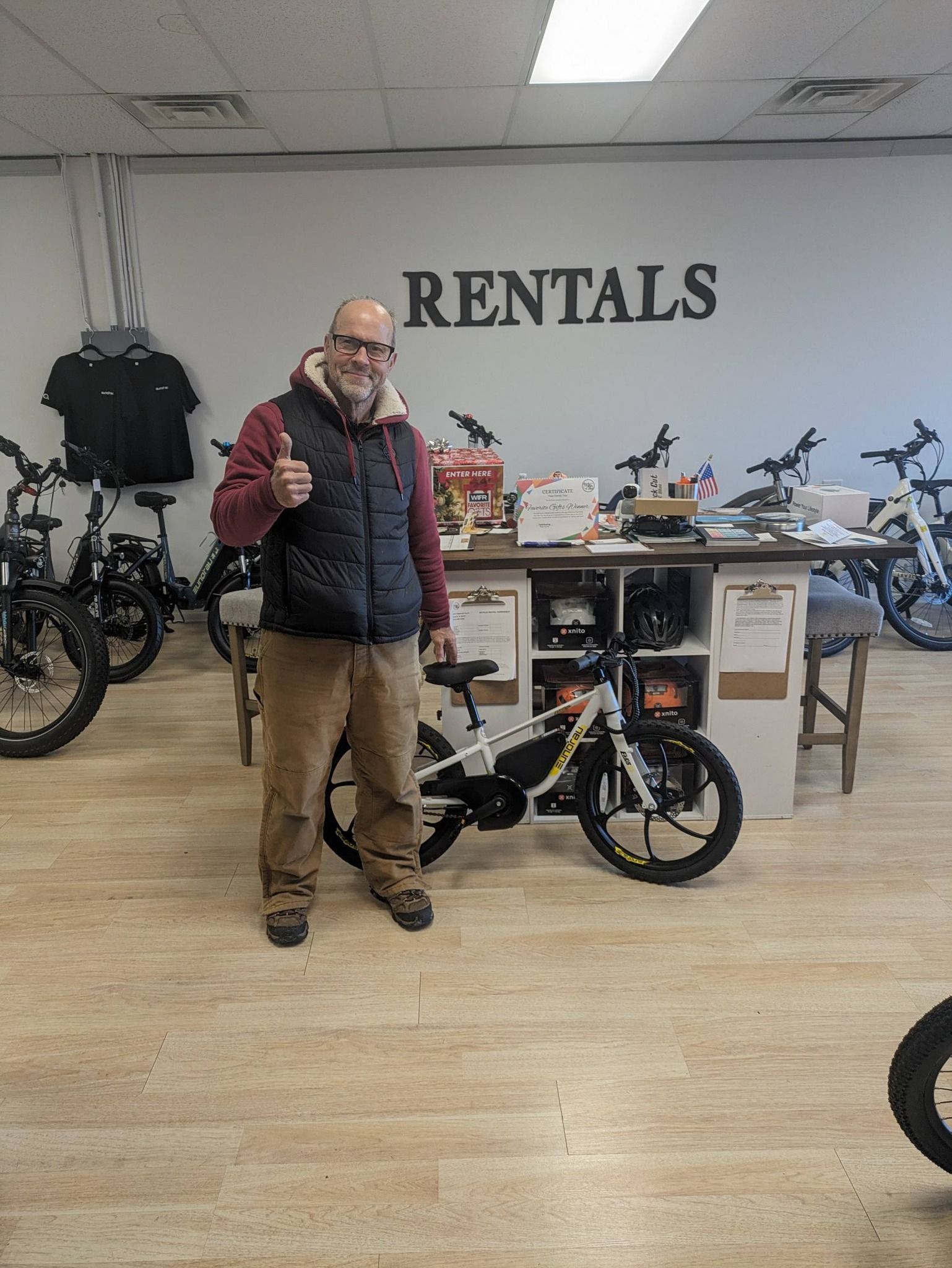 Man standing in a bike rental shop beside a black bicycle, with a “RENTALS” sign on the wall.