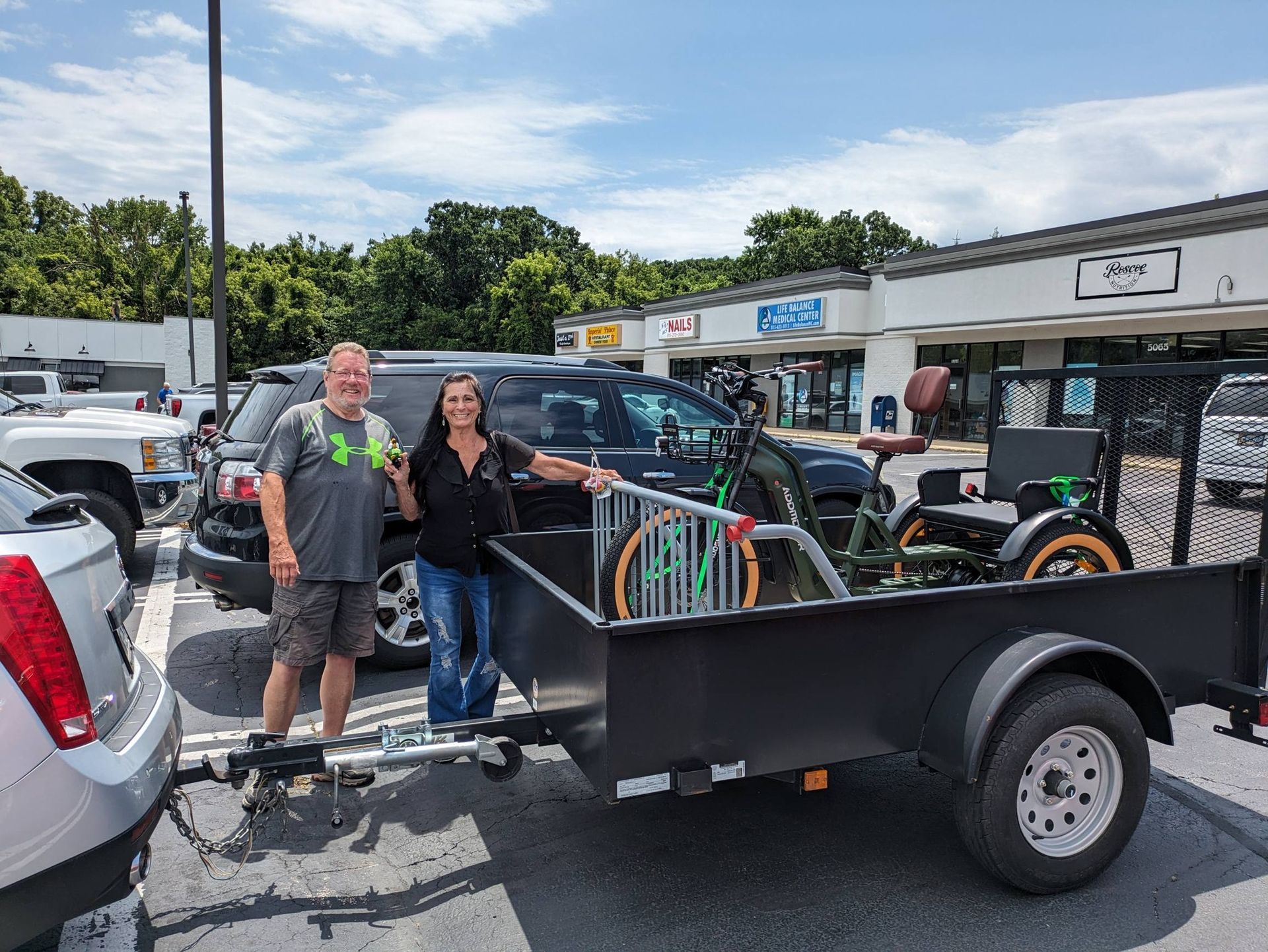 Two people standing beside a black utility trailer in a parking lot, with storefronts and parked cars behind them