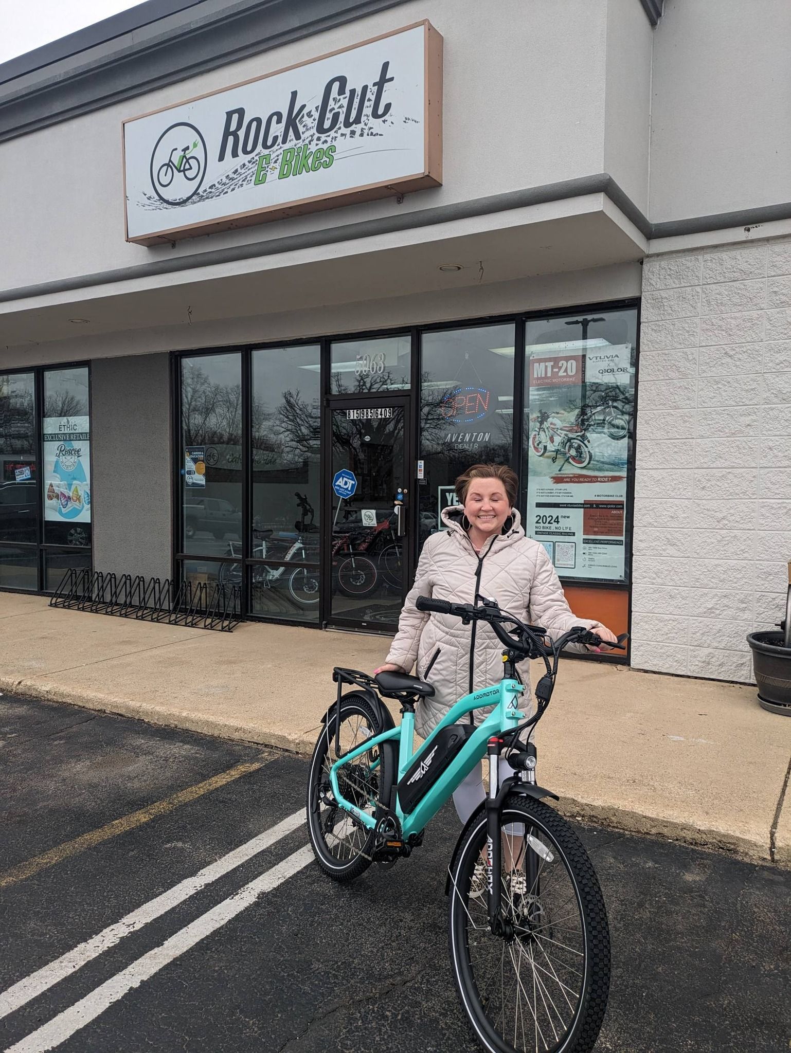 Person standing with a teal mountain bike outside a bike shop storefront.