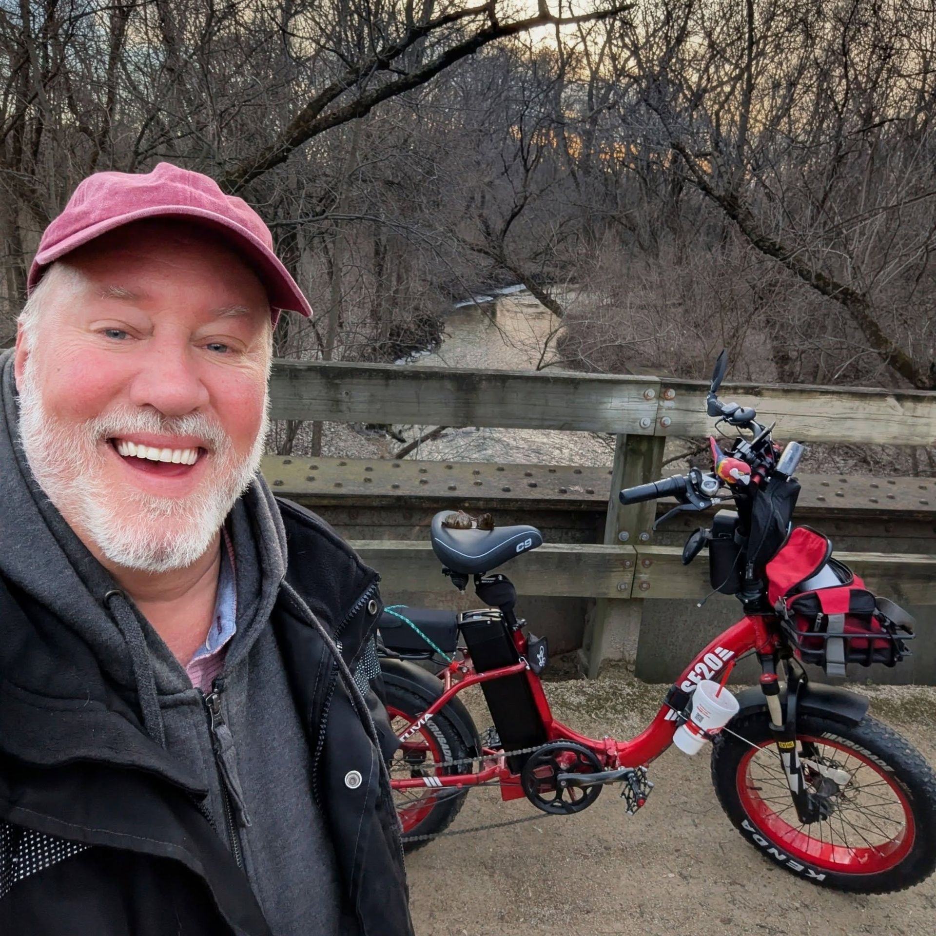 Smiling man in pink cap beside a red electric bike on a bridge by bare trees