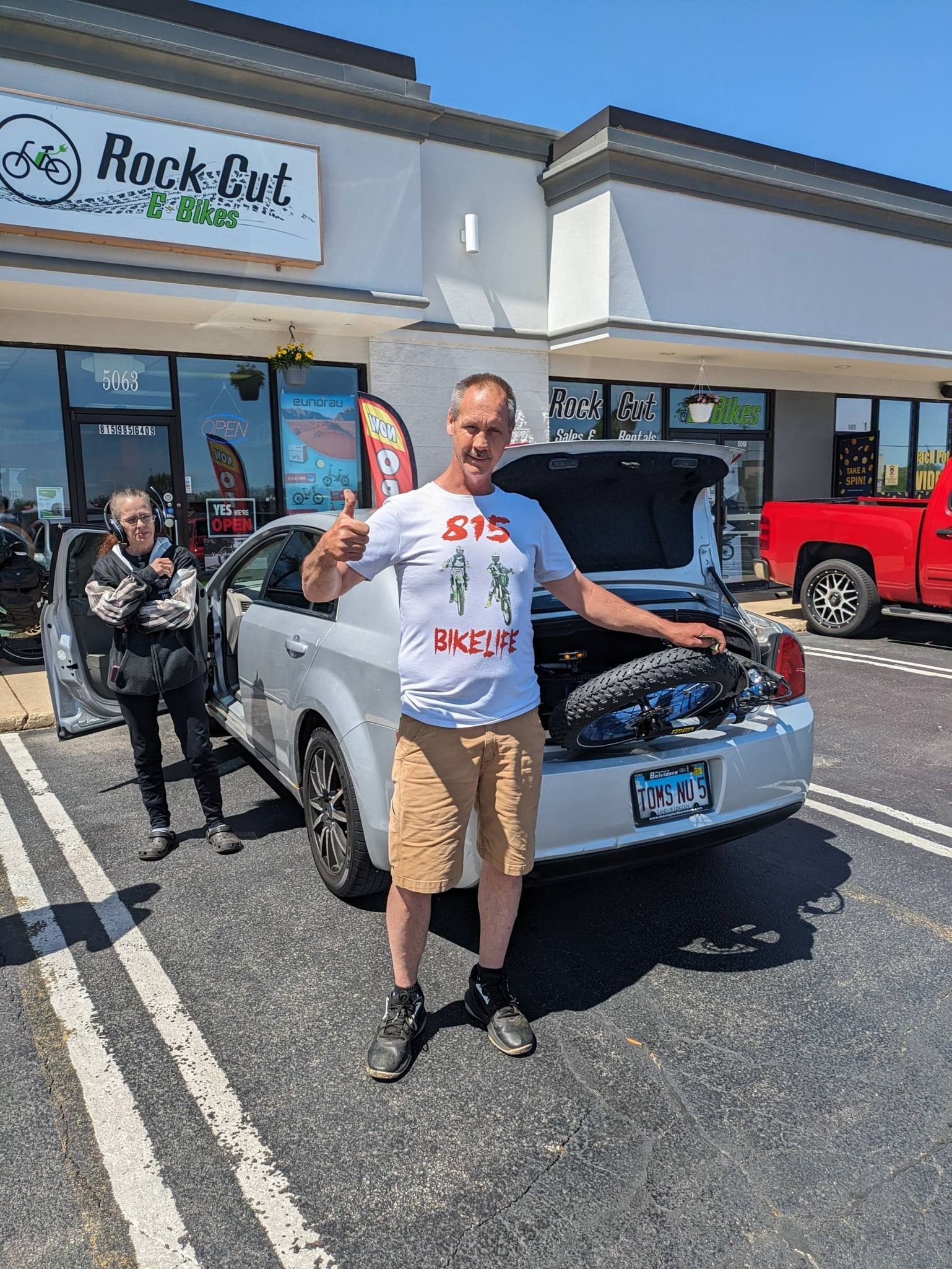 Man posing beside a silver classic car in a sunny parking lot outside a storefront