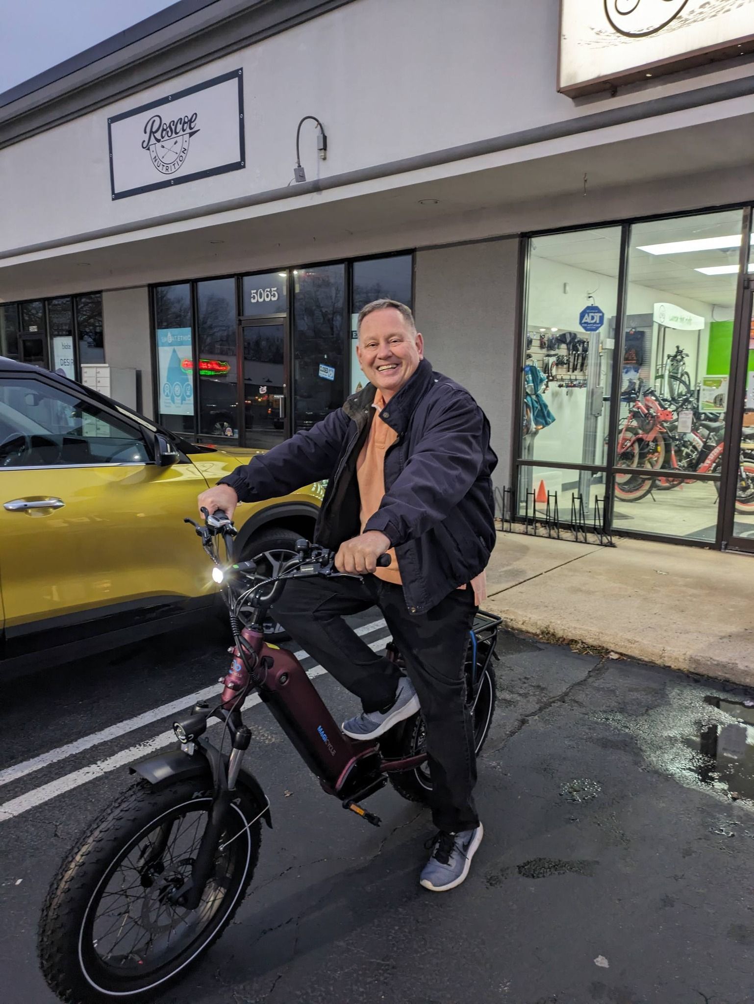Person on a black electric bike outside a storefront, smiling beside a yellow car.
