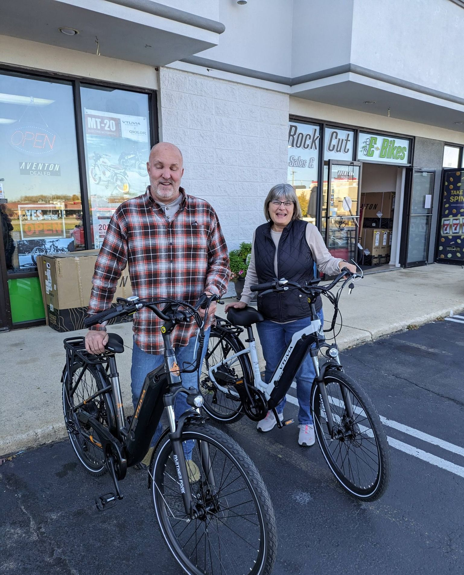 Two cyclists with bikes standing outside a storefront on a sidewalk, smiling at the camera.