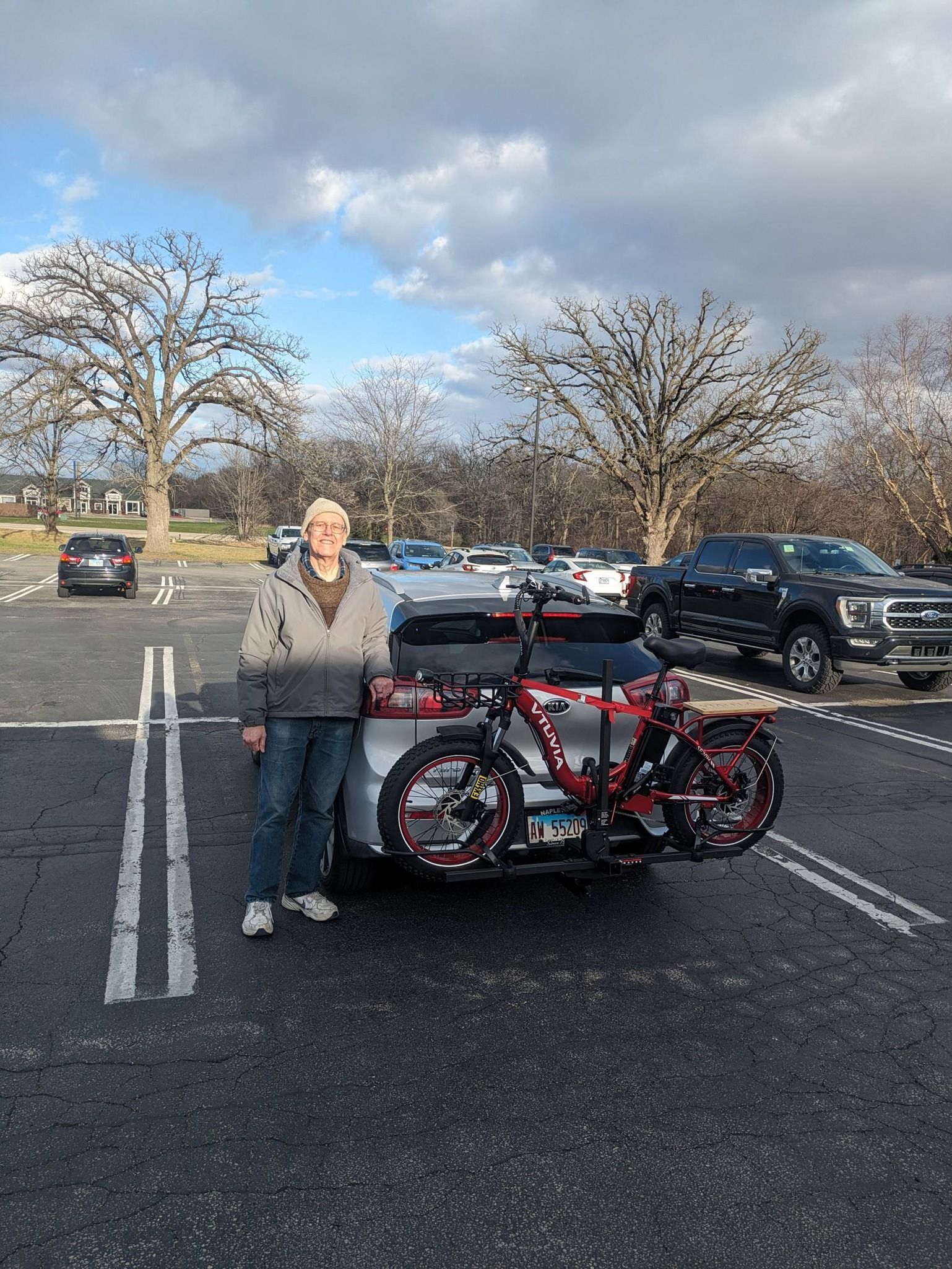 Person standing beside a red bicycle in a parking lot with parked cars and bare trees under a cloudy sky