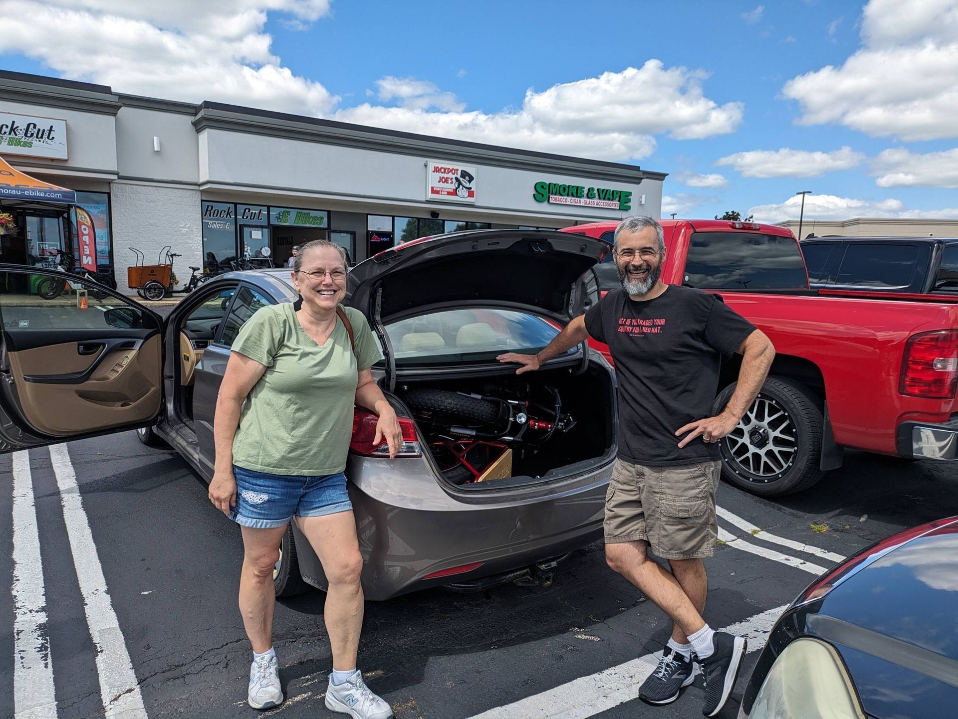 Two people posing by an open car trunk in a parking lot, with a red pickup nearby and storefronts behind.