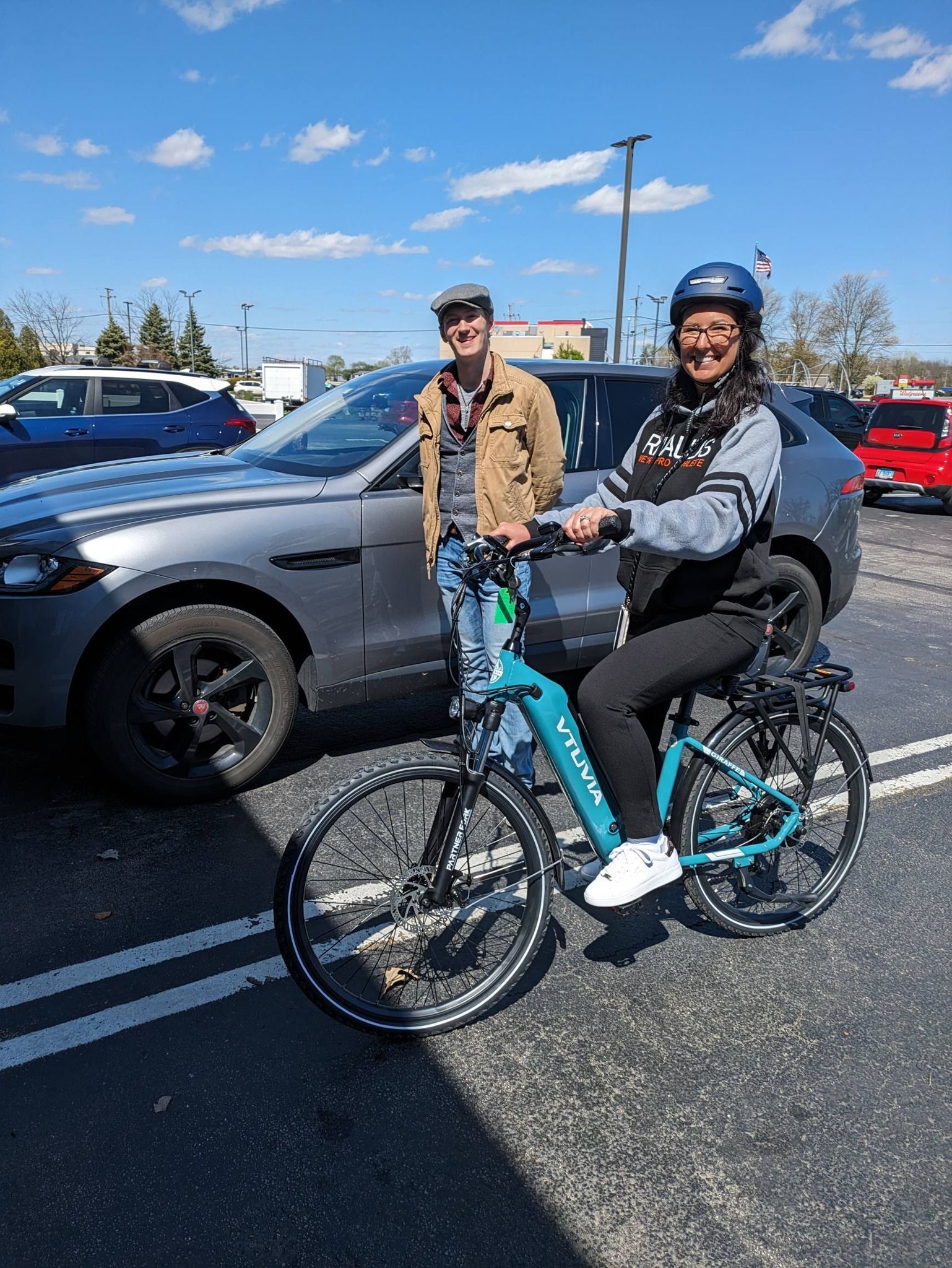 Two cyclists posing beside a gray car in a sunny parking lot, one on a turquoise bike wearing a helmet.