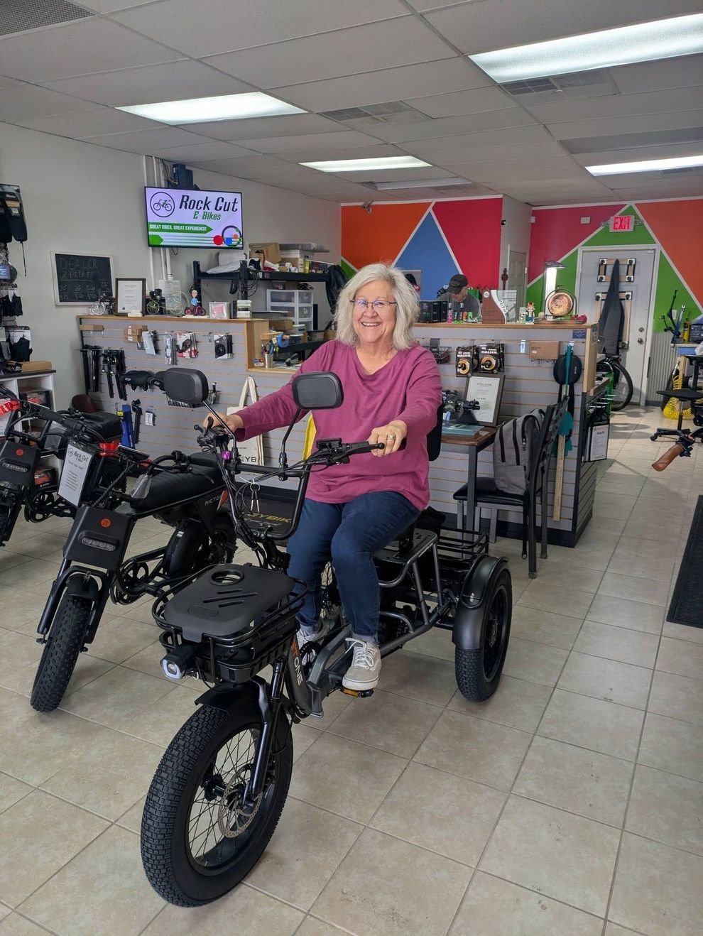 Woman seated on a black tricycle in a bike shop, smiling and pointing ahead