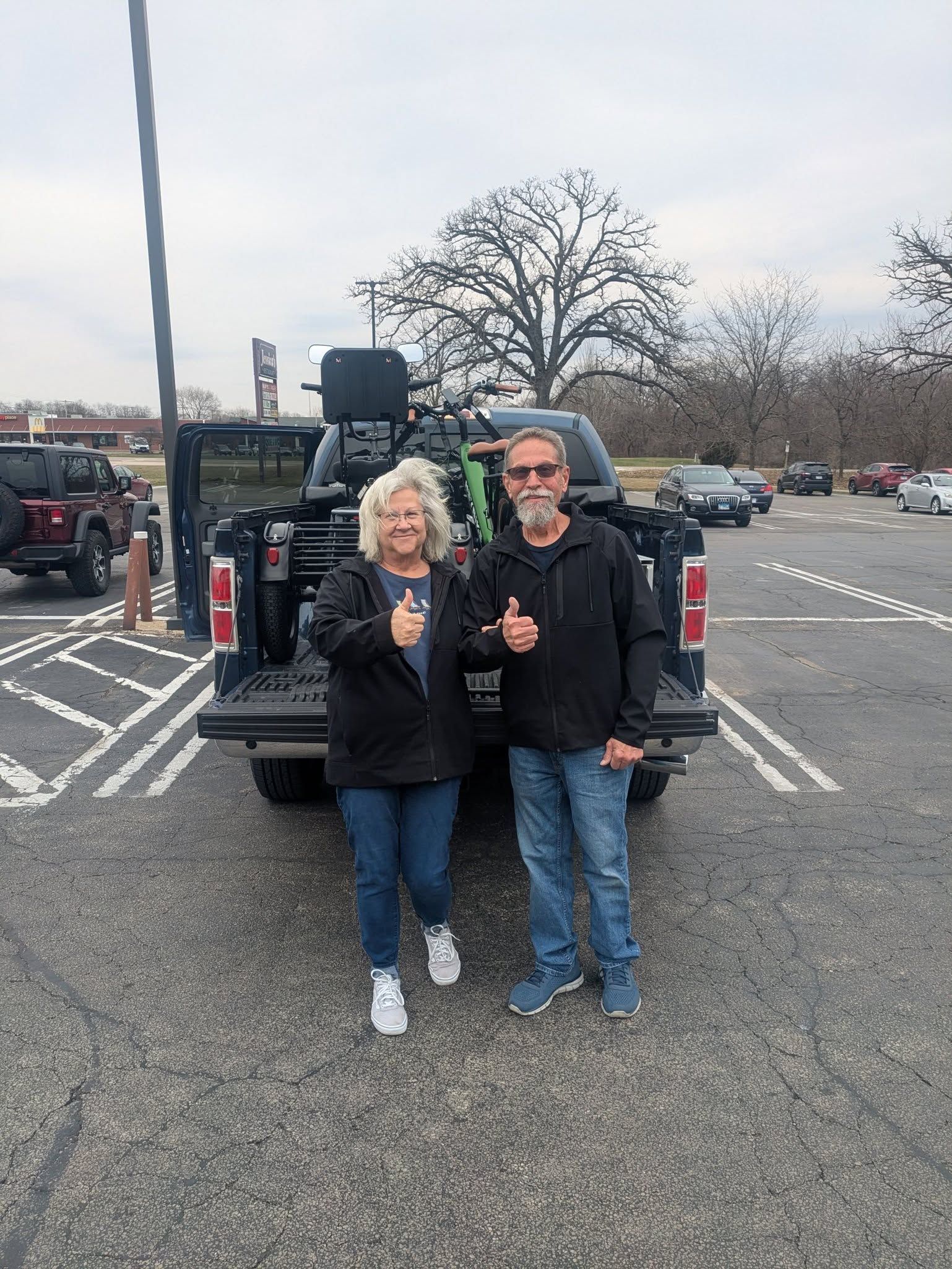 Two people standing by a parked truck in a parking lot, both giving thumbs up.