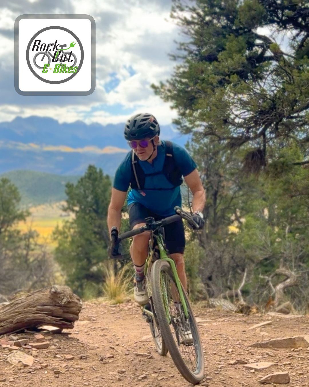 Mountain biker in blue shirt rides a dirt trail through a wooded, mountainous landscape.