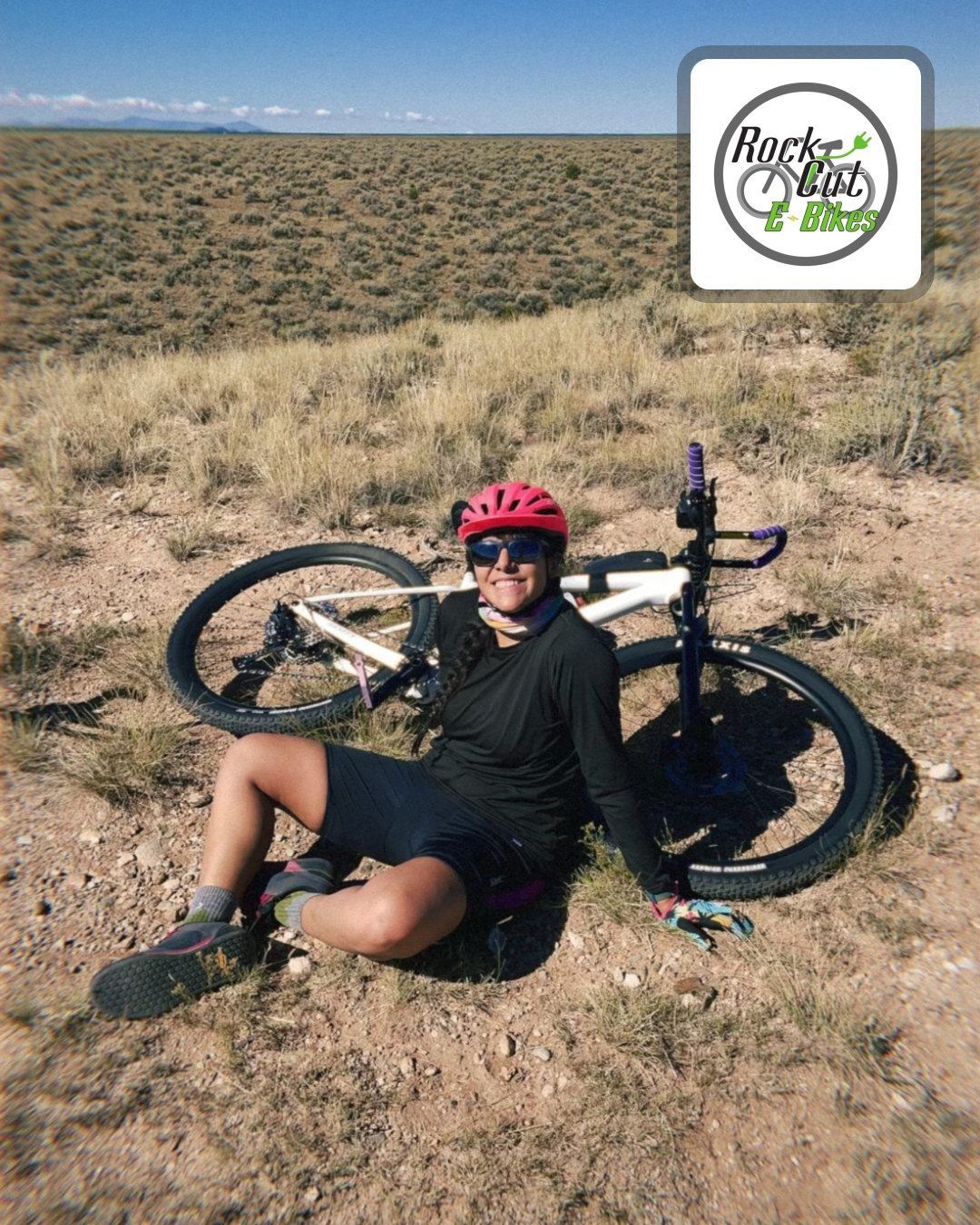 Cyclist in red helmet sitting beside a bike on a dirt trail in a desert landscape