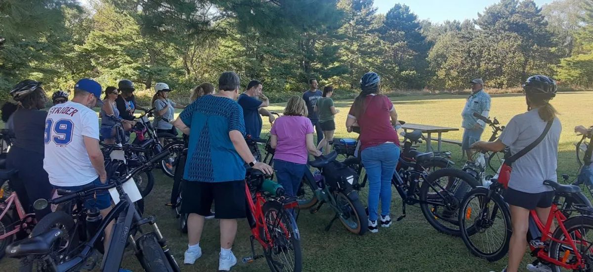 People with bicycles gathered outdoors on a sunny grassy hill, facing away toward the view