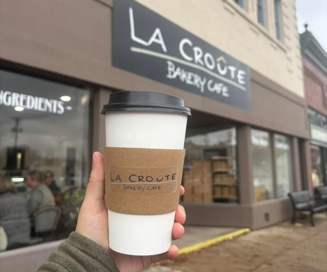 Hand holding a takeaway coffee cup in front of La Croûte Bakery Café storefront