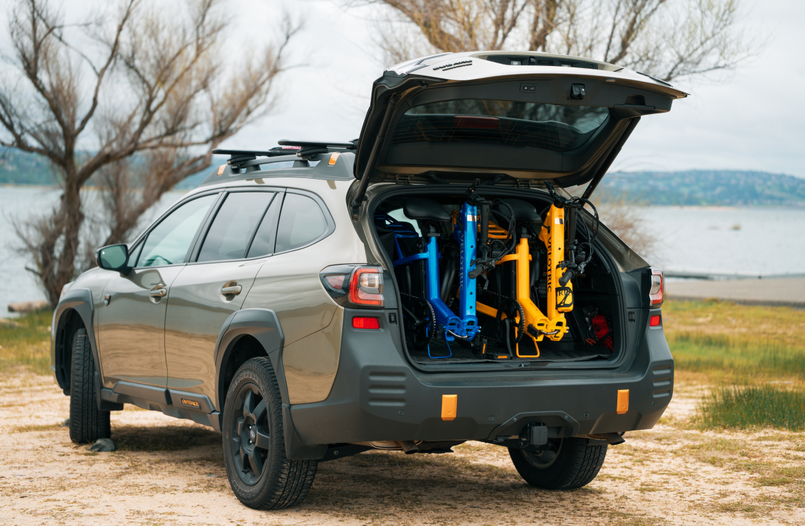 Gray SUV with open rear hatch revealing colorful bikes at a lakeside, with leafless trees in the background