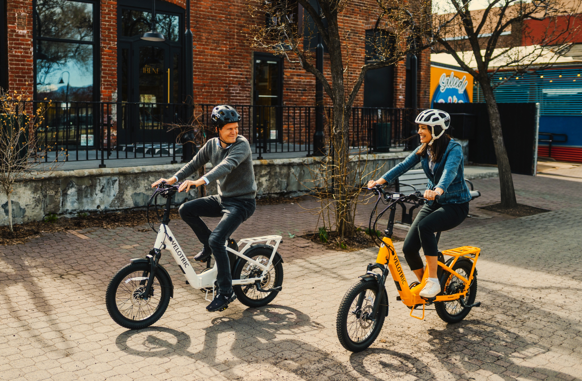 Two cyclists on electric bikes riding past brick buildings on a sunny street