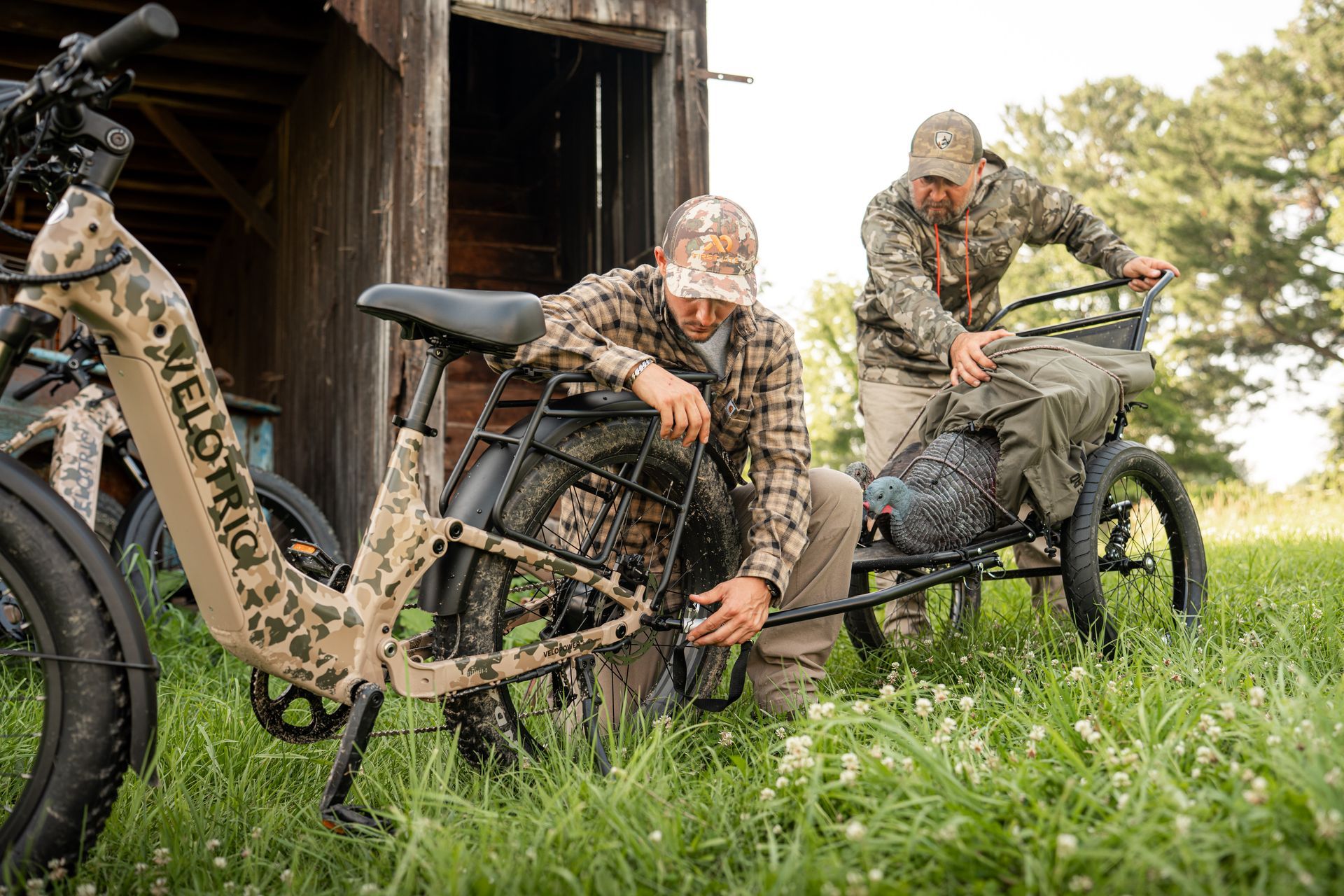 Two camouflaged hunters with an electric hunting bike beside a wooden shed in a grassy field