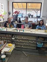 Three people stand behind a cafe counter with pastries and plants in a bright storefront