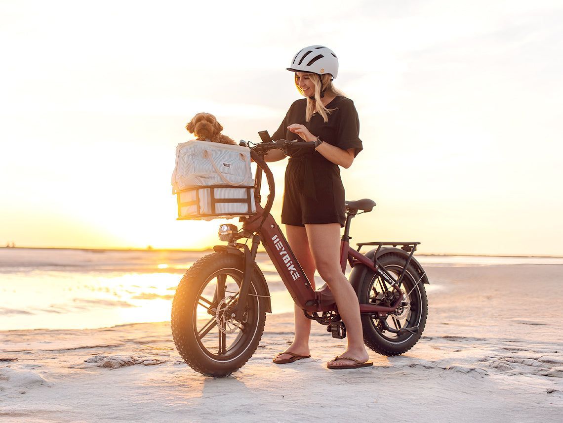 Woman with a helmet standing beside an e-bike on a sandy beach at sunset, holding a phone.
