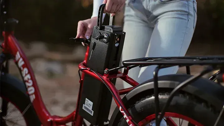 Person holds a red electric bicycle with a black battery pack attached to the frame.