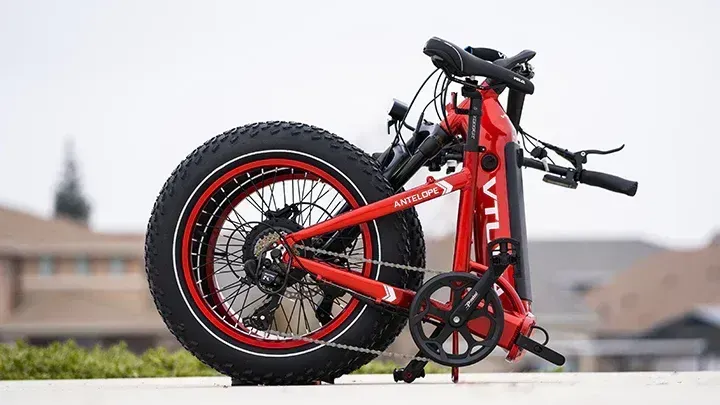 Red folding bicycle with fat tires and black accents, parked outdoors on a ledge against a blurred building background