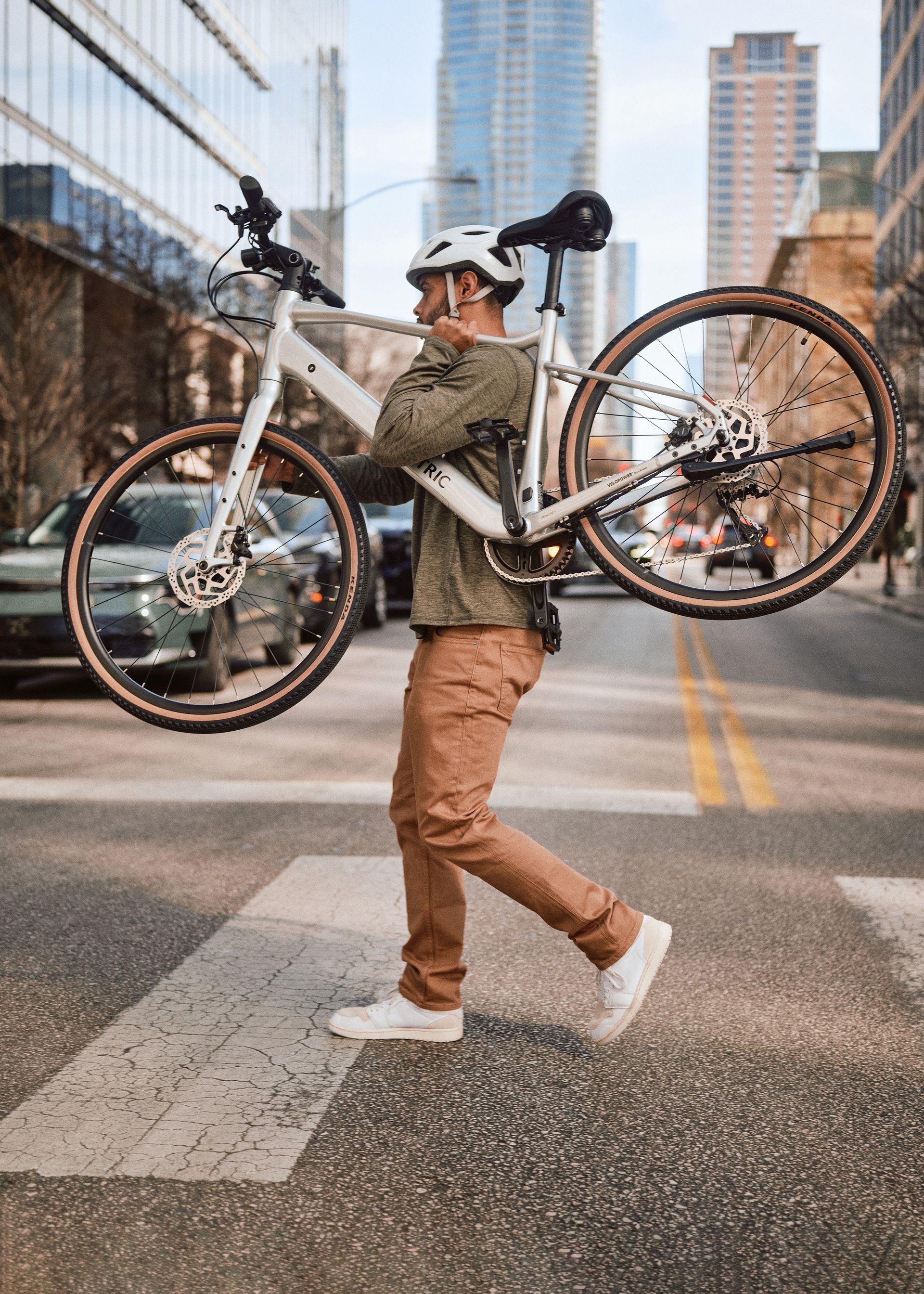 Person carrying a bicycle across a city street with tall buildings in the background