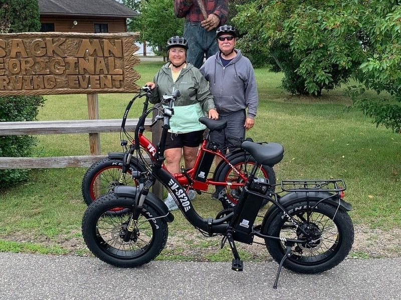 Two cyclists posing with their bikes in front of a wooden sign and grassy park backdrop