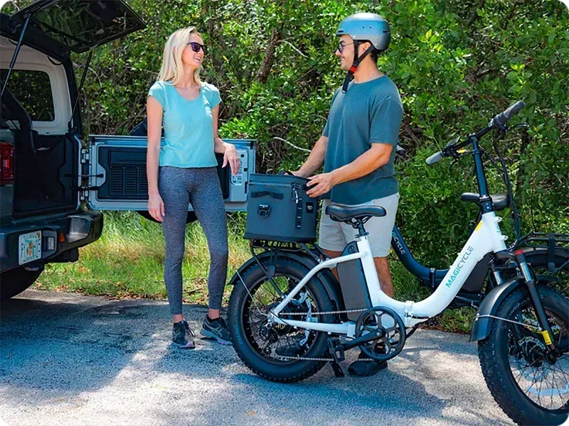 Two people chatting beside a white electric bike and car trunk on a wooded roadside