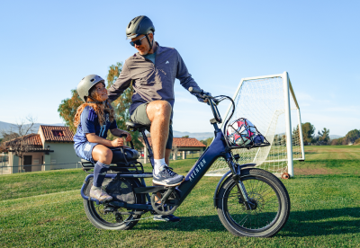 Two people riding a cargo bike on a grassy field near a soccer goal, smiling in helmets