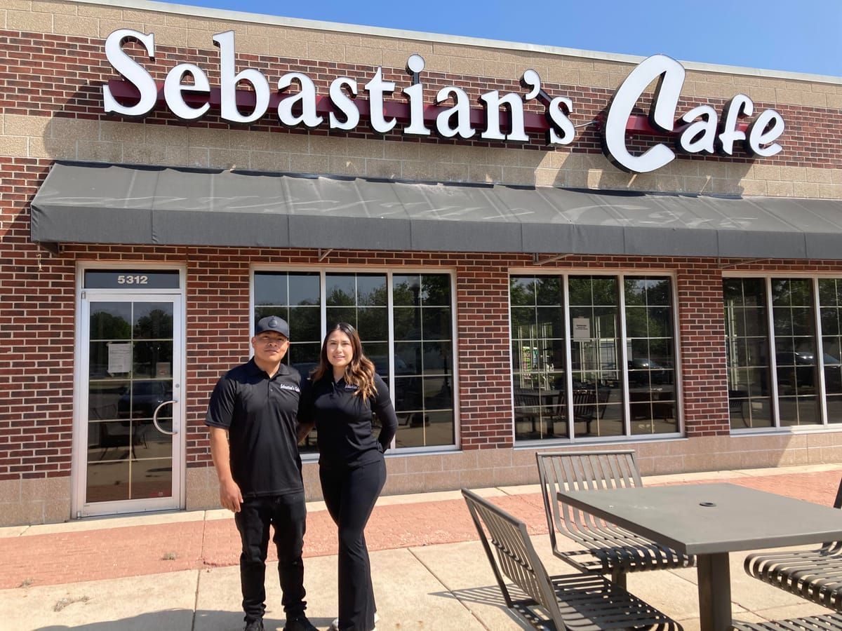 Two people standing outside Sebastian’s Cafe in front of a brick storefront.