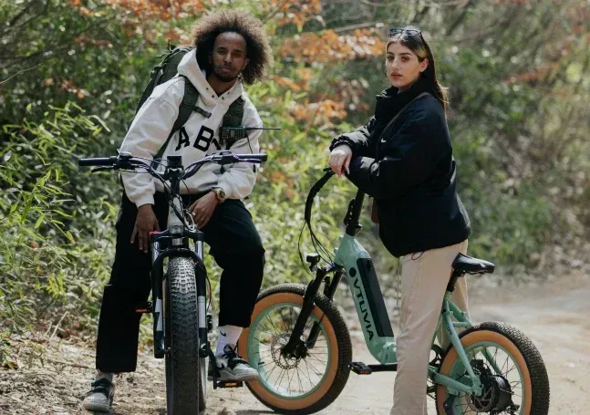 Two cyclists pause on a wooded trail, standing with their bikes in autumn foliage.