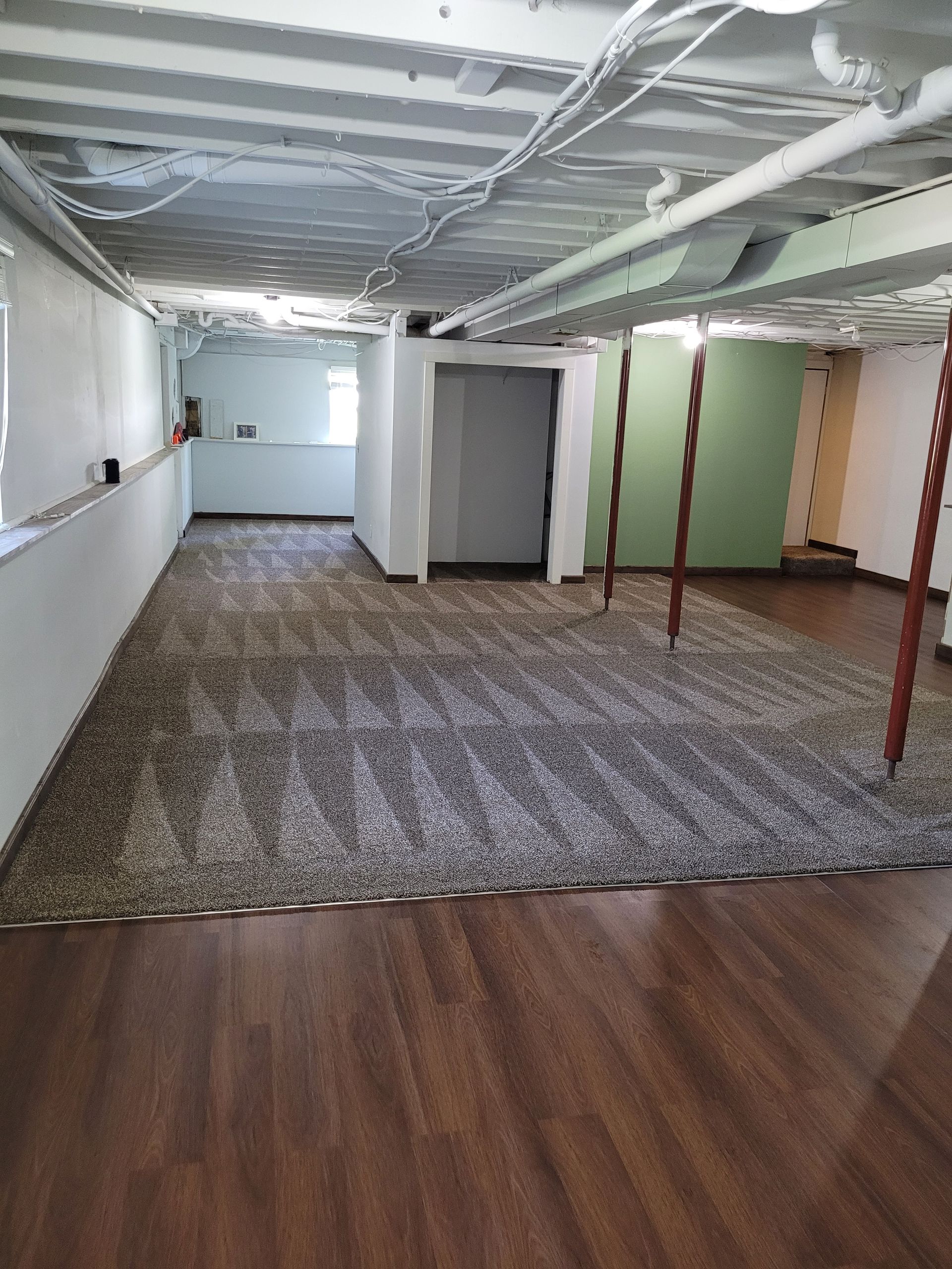 Finished basement with wood floor, carpeted area, support beams, and utility pipes on ceiling.