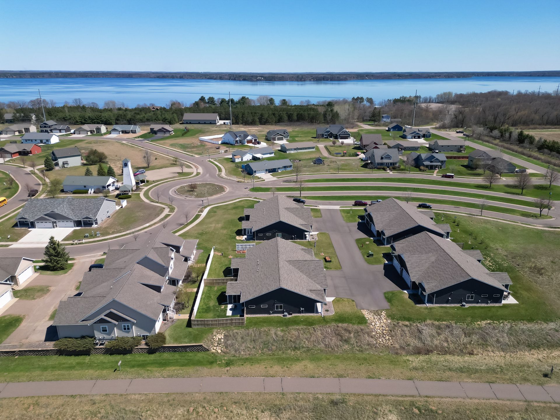 Aerial view of a residential area with gray houses, a roundabout, and a lake in the background under a blue sky.