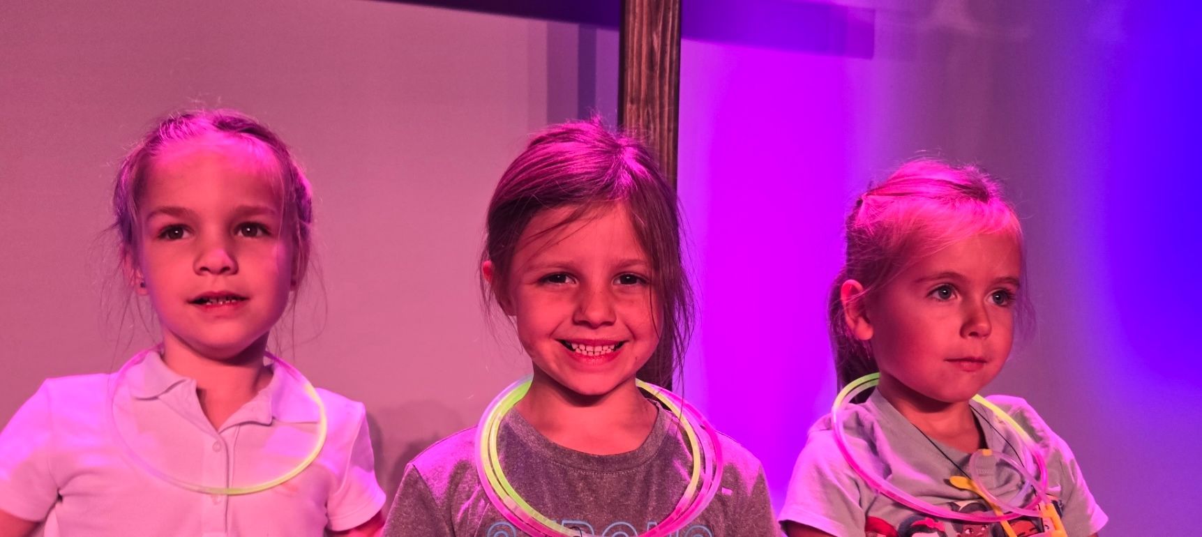 Three little girls wearing neon necklaces are standing next to each other on a stage.