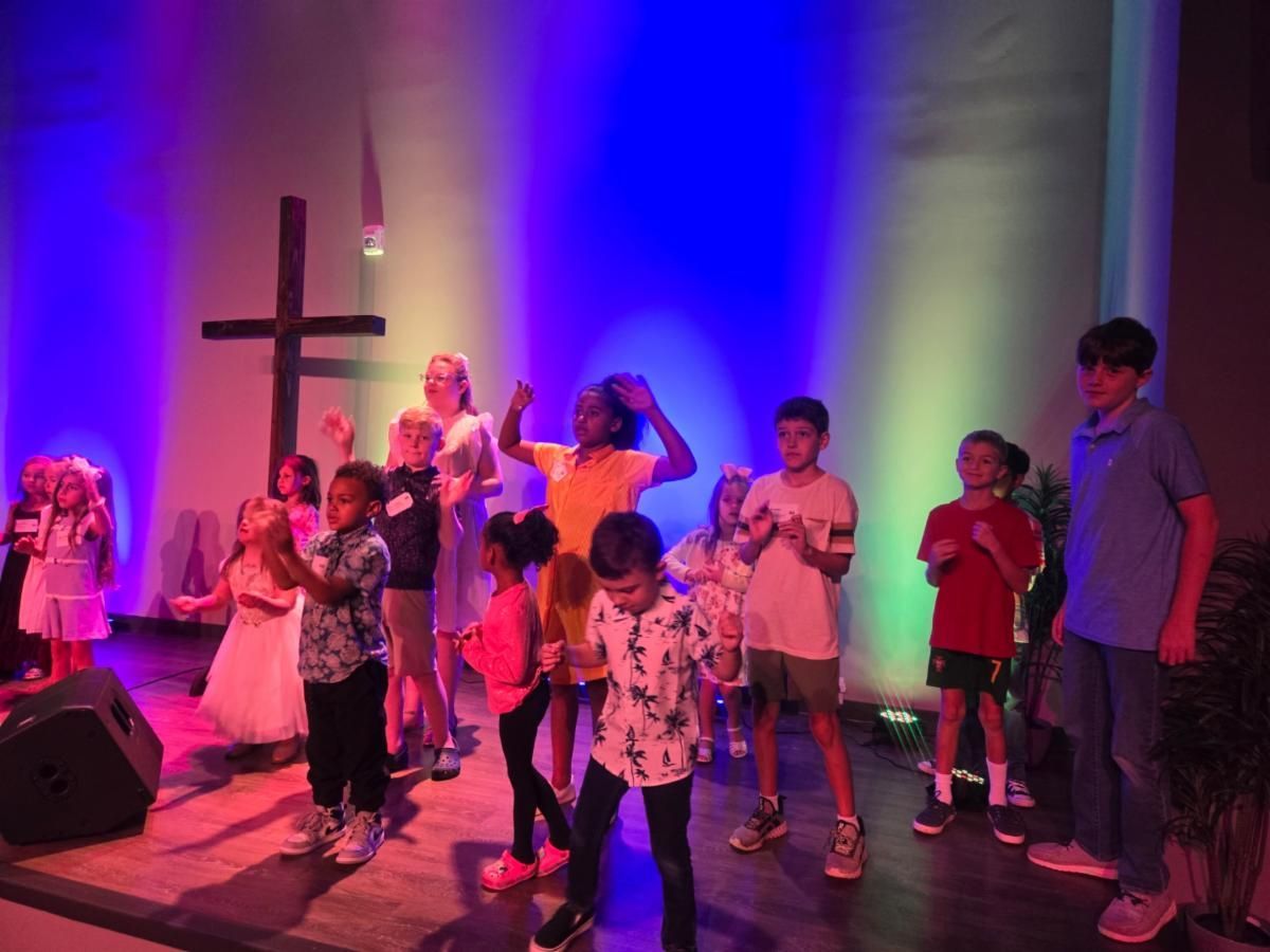 A group of children are dancing on a stage in front of a cross.