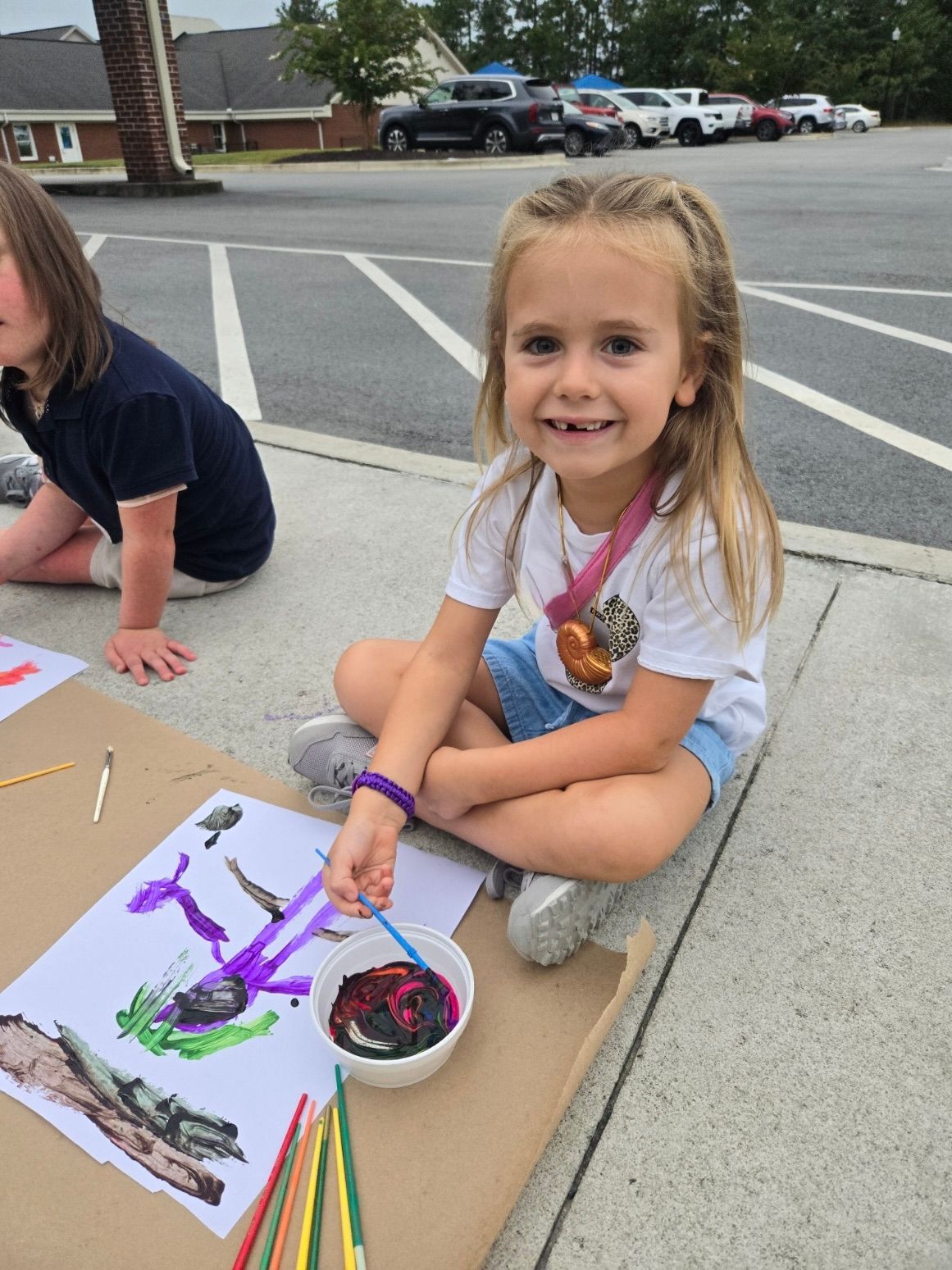 A little girl is sitting on the sidewalk painting a picture.